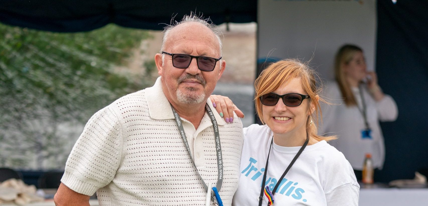 Trivallis Housing Landlord Wales A man and a woman standing outdoors at an event. the man is bald with glasses and wears a white sweater and denim shorts. the woman, in glasses, sports a logo t-shirt and denim shorts. both smile at the camera.