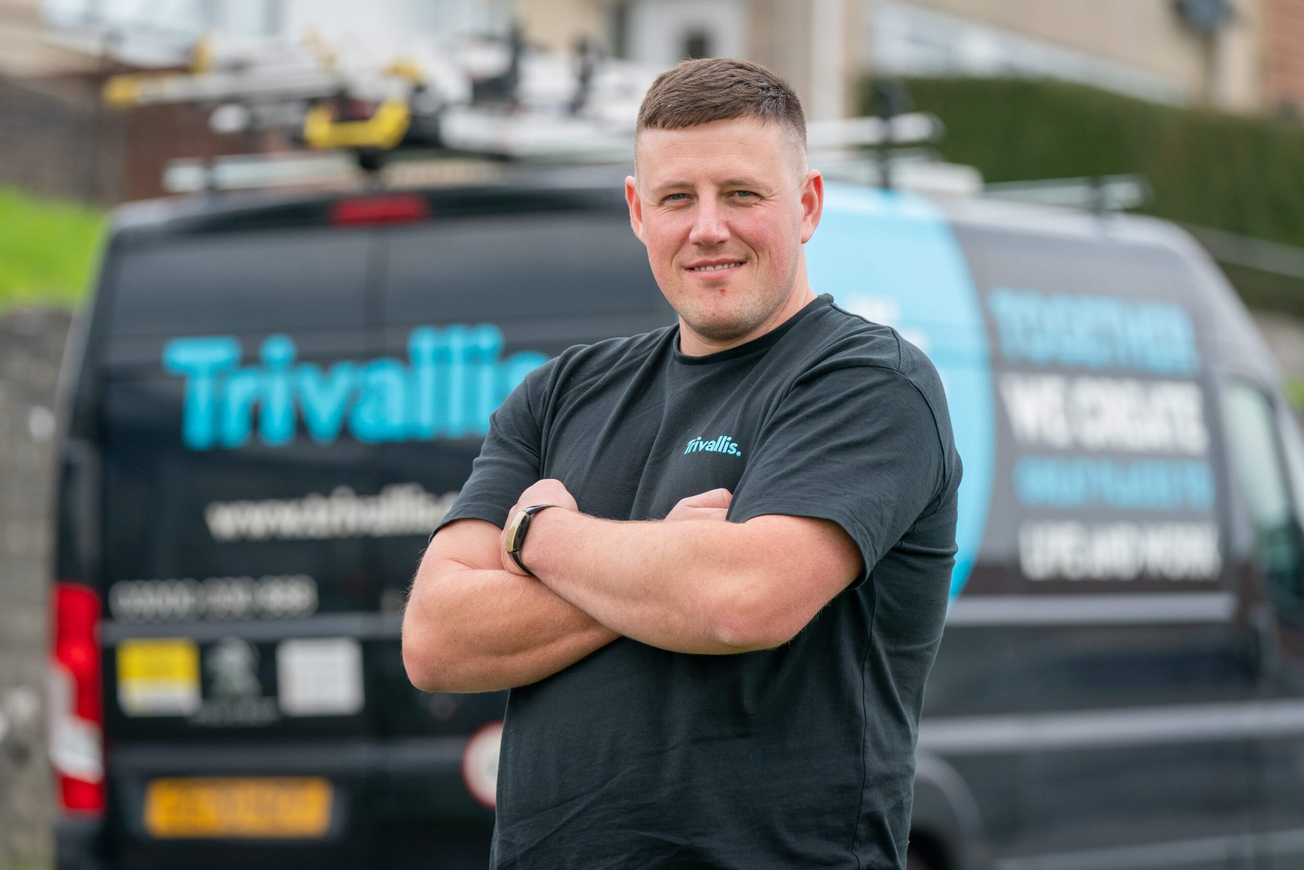 A man with crossed arms smiling in front of a service van with "trivallis" branding.