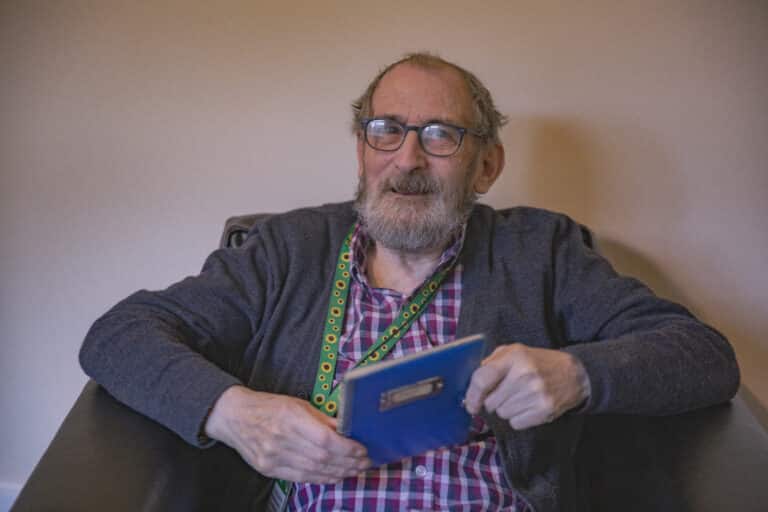 Trivallis Housing Landlord Wales An elderly man wearing eyeglasses, a plaid shirt, and a cardigan, sitting and smiling slightly while holding a blue book about Trivallis housing.