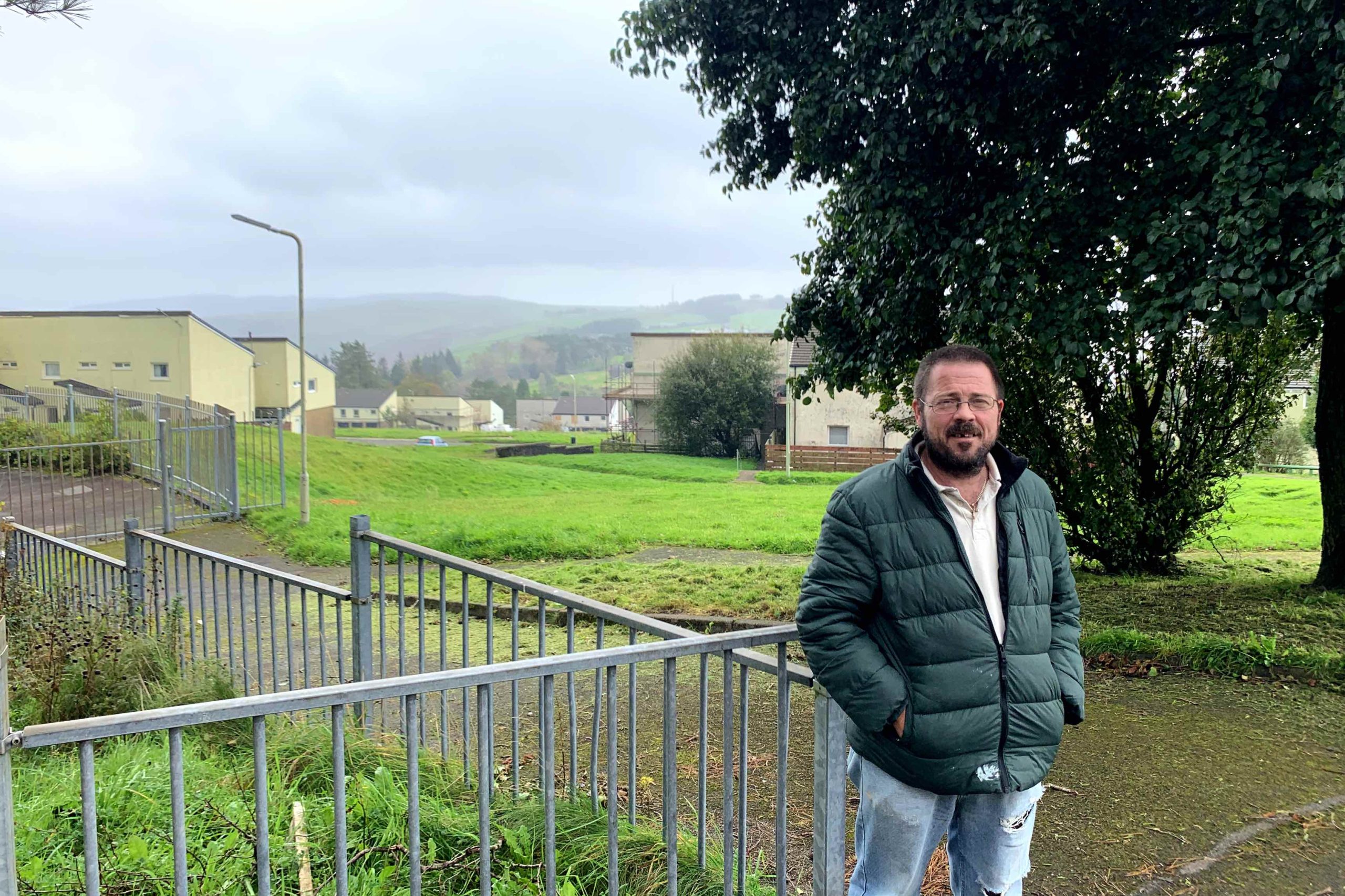 Trivallis Housing Landlord Wales A man stands in front of a metal fence with a Trivallis housing area and green hills in the background, under a cloudy sky.