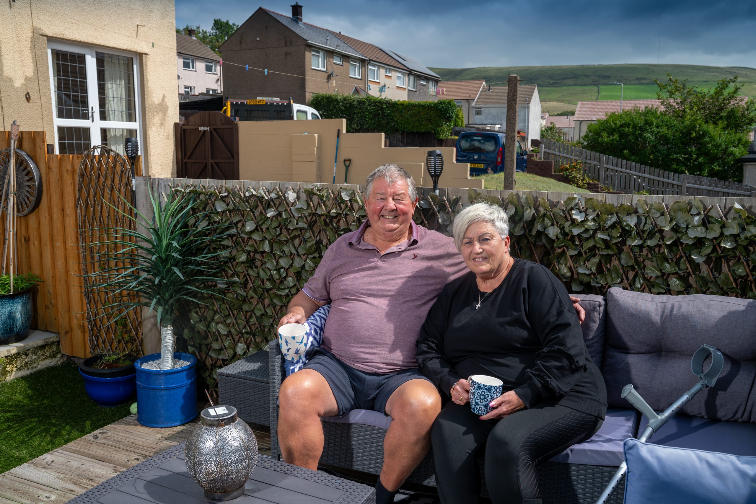 A smiling older couple sitting together on outdoor furniture in a backyard with houses and hills in the background.