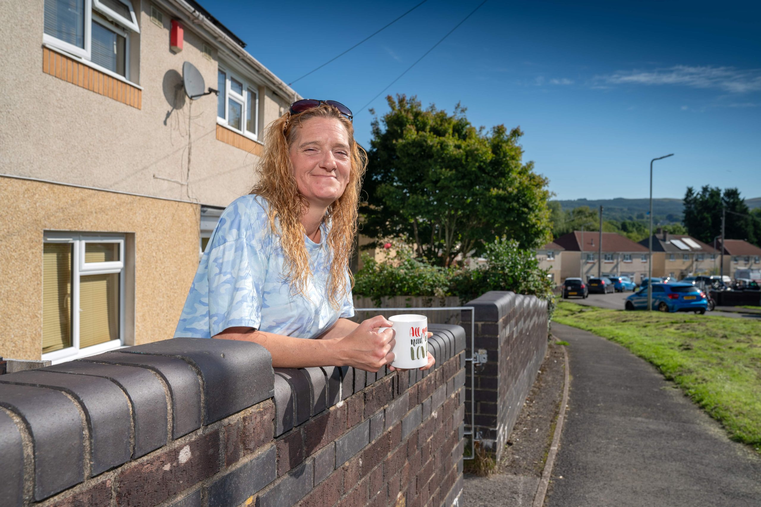 Trivallis Housing Landlord Wales A woman with long hair leaning on a fence, holding a mug, with a Trivallis residential street in the background.