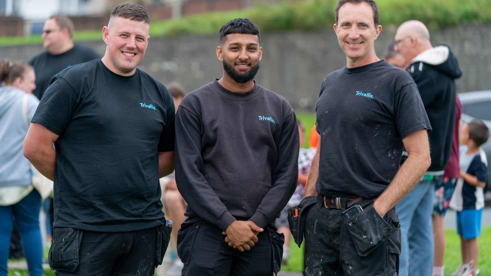 Three smiling men in black work shirts and trousers standing together outdoors.