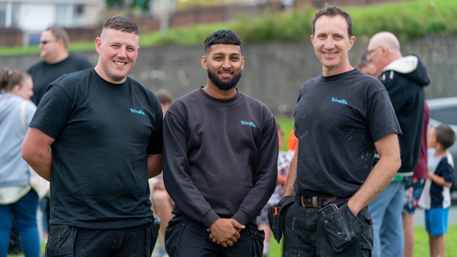 Three smiling men in black work shirts and trousers standing together outdoors.