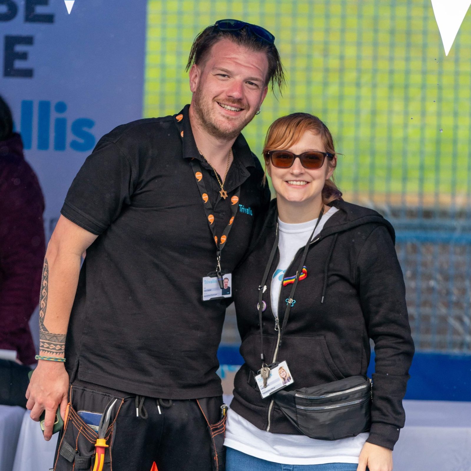 Trivallis Housing Landlord Wales Two individuals, a man and a woman, pose for a photo together, smiling. The man is wearing a black shirt with orange accents and a tool belt, while the woman is in a black hoodie
