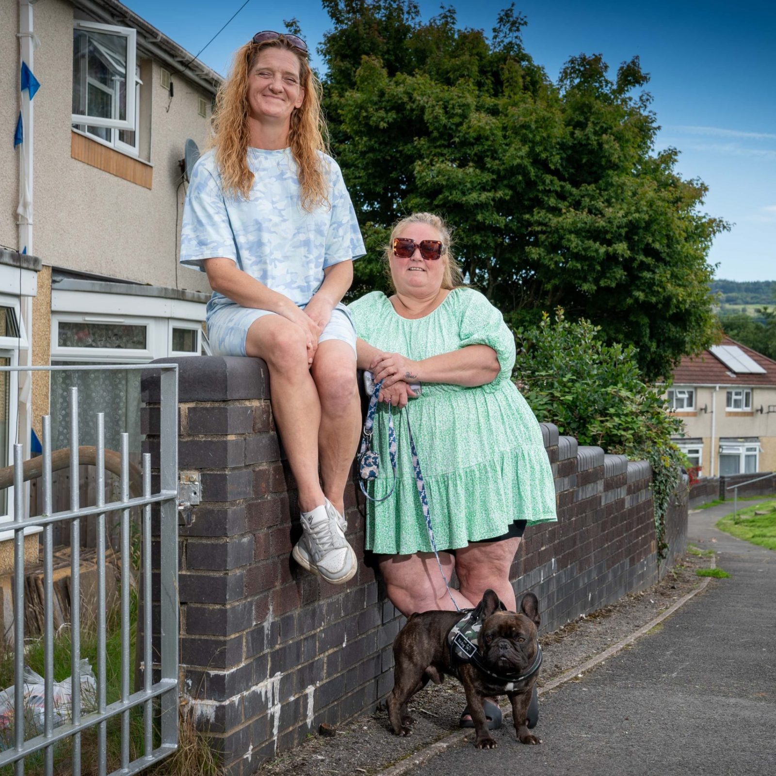 Trivallis Housing Landlord Wales Two women sitting on a low wall outside a row of Trivallis houses, accompanied by a small black dog on a leash.