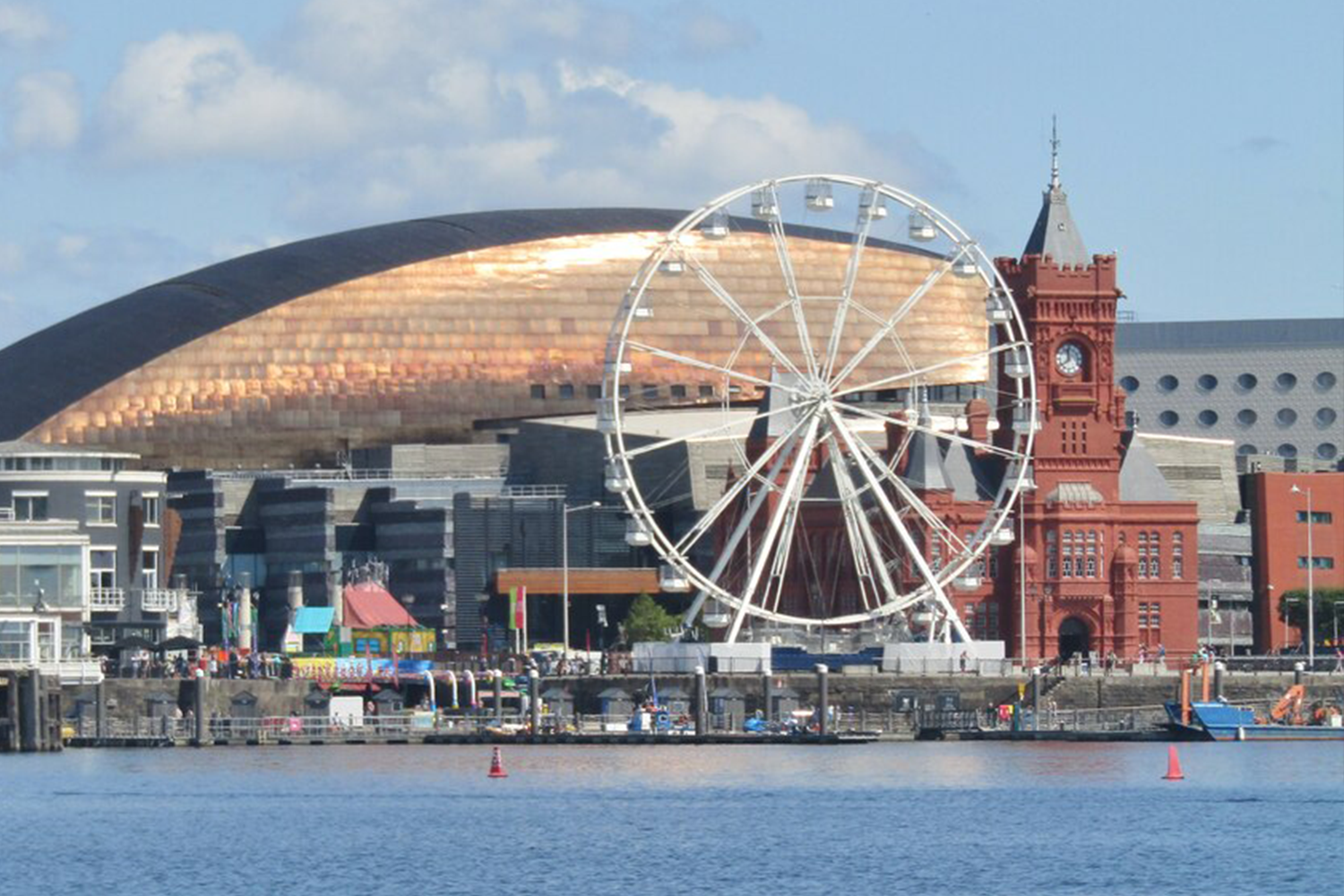 Trivallis Housing Landlord Wales A ferris wheel and a distinctive red brick housing building stand in front of an arena with a reflective dome, along a waterfront.