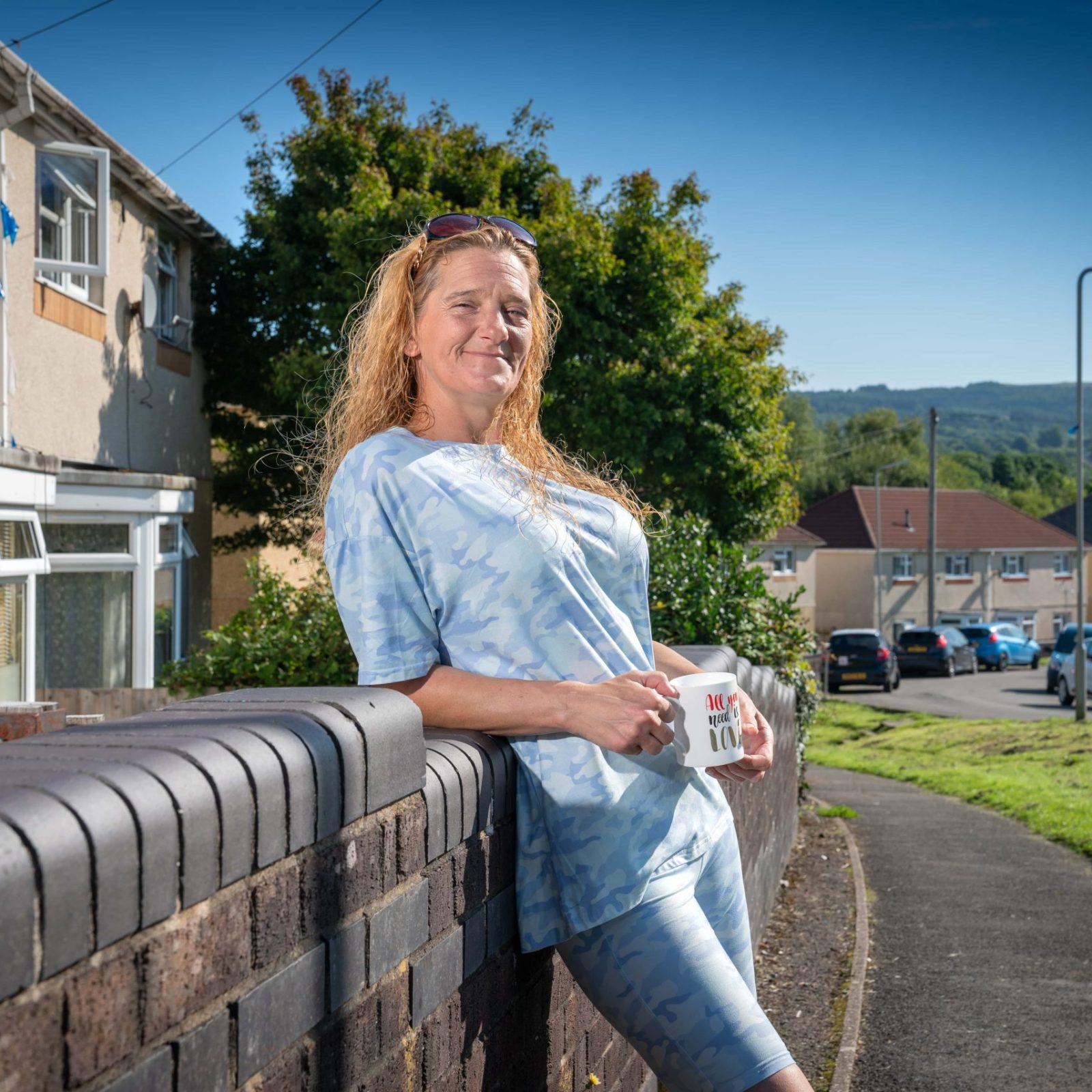 Trivallis Housing Landlord Wales A smiling woman holding a mug stands leaning on a brick wall on a sunny RCT suburban street.