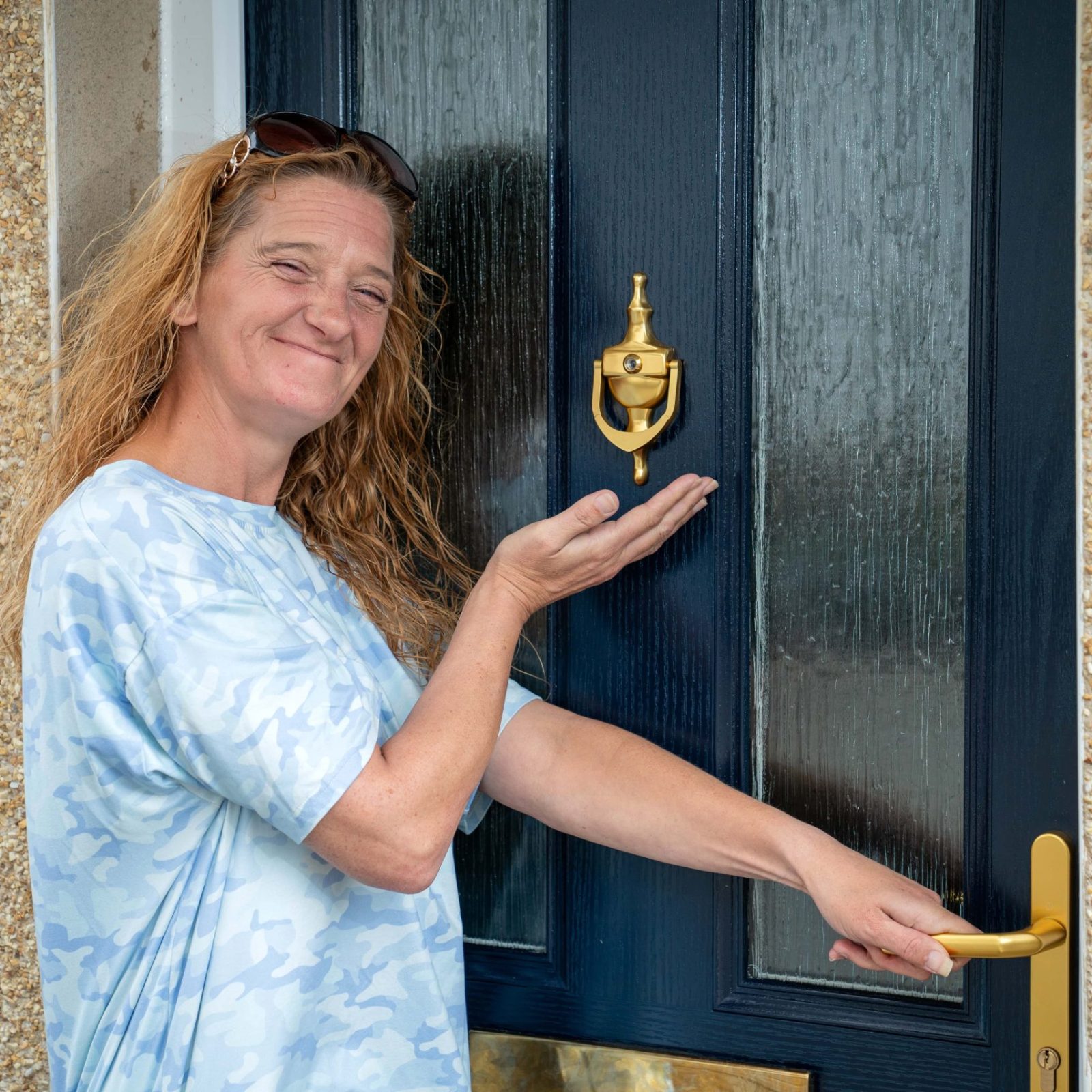 Trivallis Housing Landlord Wales A woman with sunglasses on her head is smiling while presenting or gesturing toward a black Trivallis housing door with a brass knocker and handle.