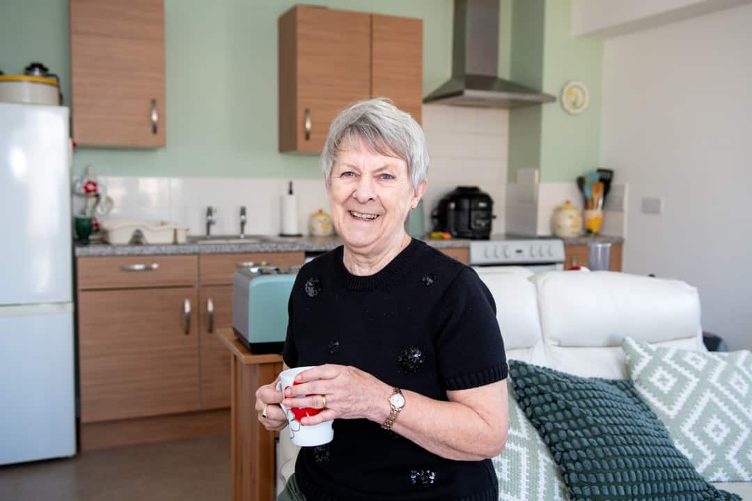 Trivallis Housing Landlord Wales An elderly woman smiling at the camera, holding a mug in a well-lit kitchen with modern appliances in Trivallis housing.