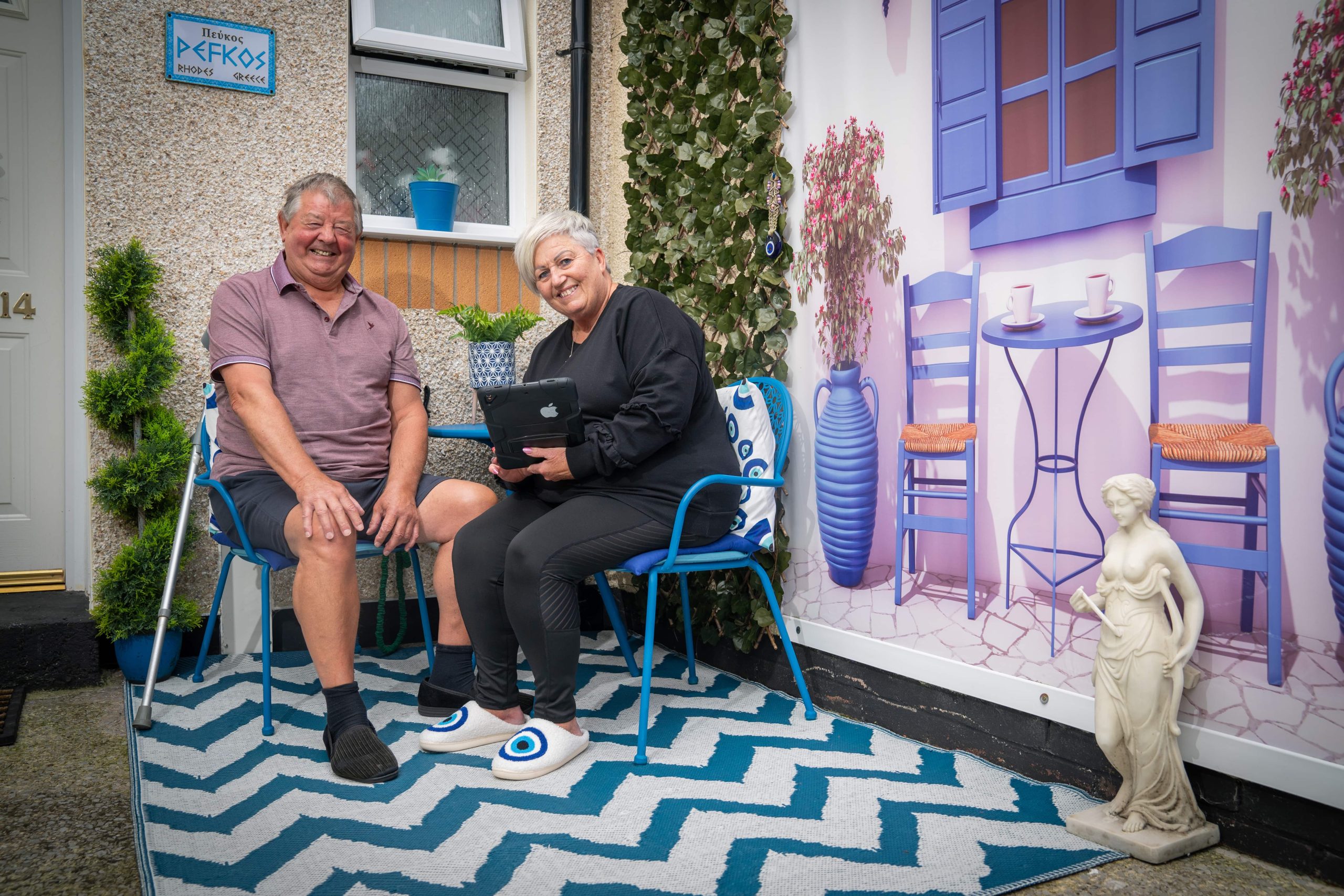 Trivallis Housing Landlord Wales A man and a woman sit outside a house with a decorative facade featuring a painted scene of a cafe, complete with a statue and a patterned rug, part of Trivallis' RCT housing