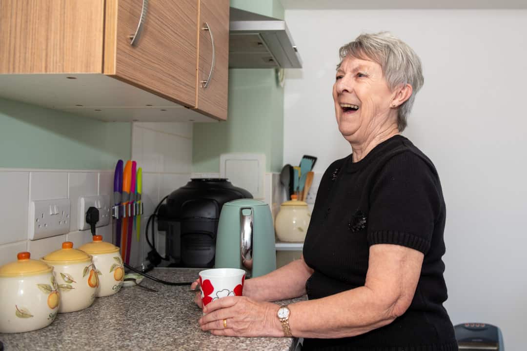 Trivallis Housing Landlord Wales An elderly woman laughing in a Trivallis kitchen, holding a red mug, with kitchen appliances and utensils in the background.