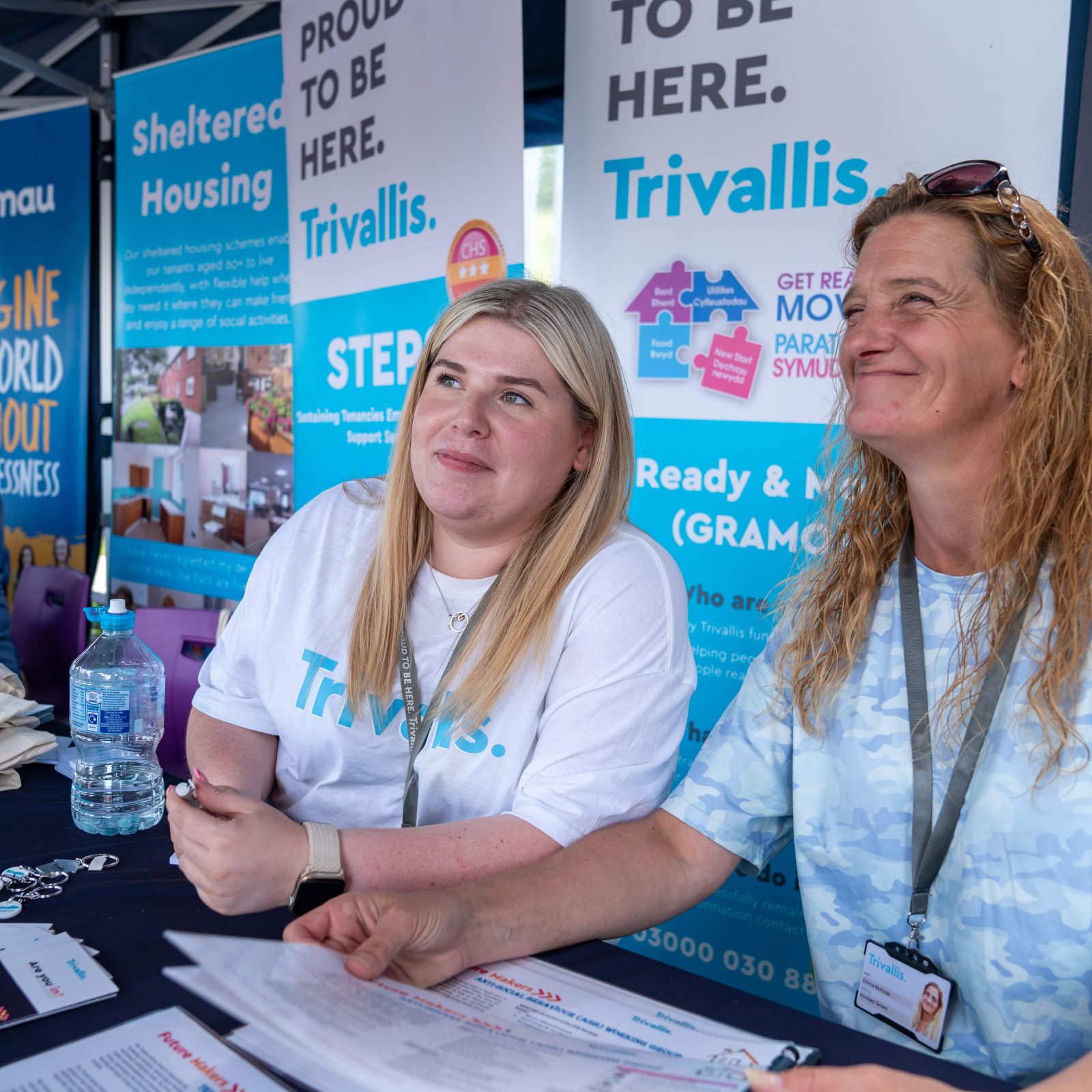 Trivallis Housing Landlord Wales Two women sitting at a booth with banners and promotional materials for Trivallis, a housing organization based in RCT, at a public event.