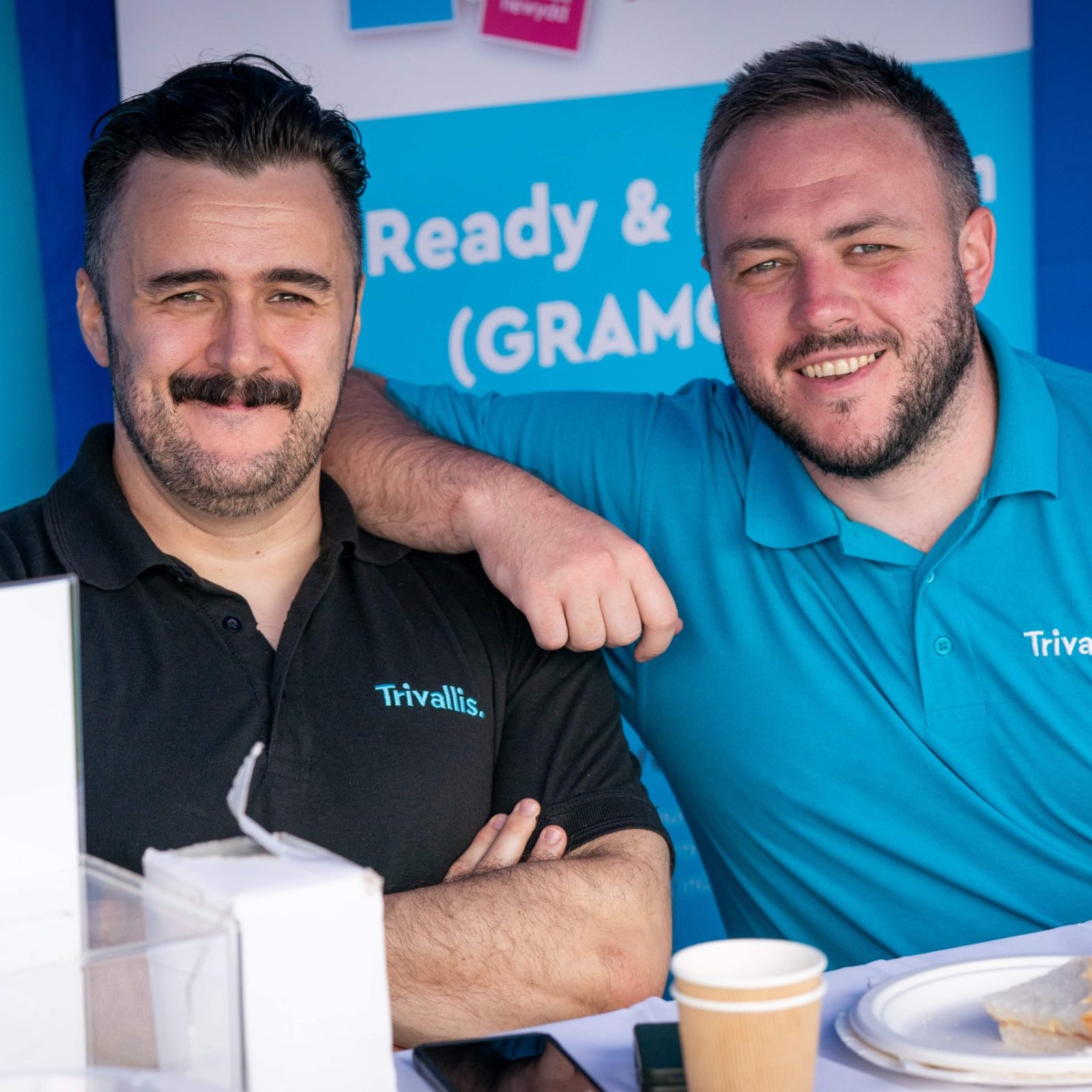 Trivallis Housing Landlord Wales Two men in blue polo shirts, bearing the logo 'Trivallis,' are smiling and posing together at a Trivallis housing promotional booth with a paper cup and a piece of cake on the table