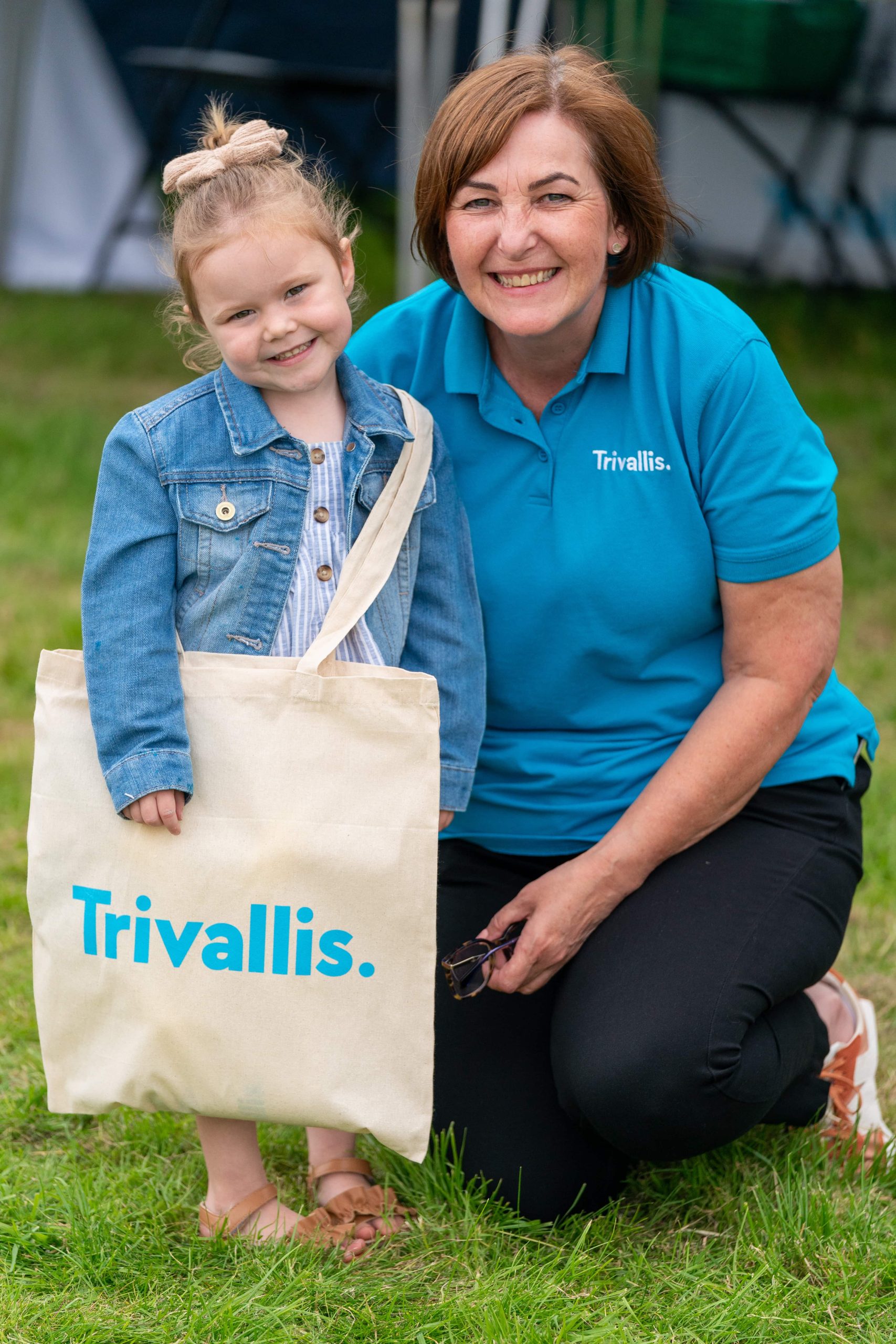 Trivallis Housing Landlord Wales A young girl with a ponytail and a woman in a blue Trivallis shirt are smiling at the camera while kneeling on grass, holding a tote bag with the RCT housing logo.