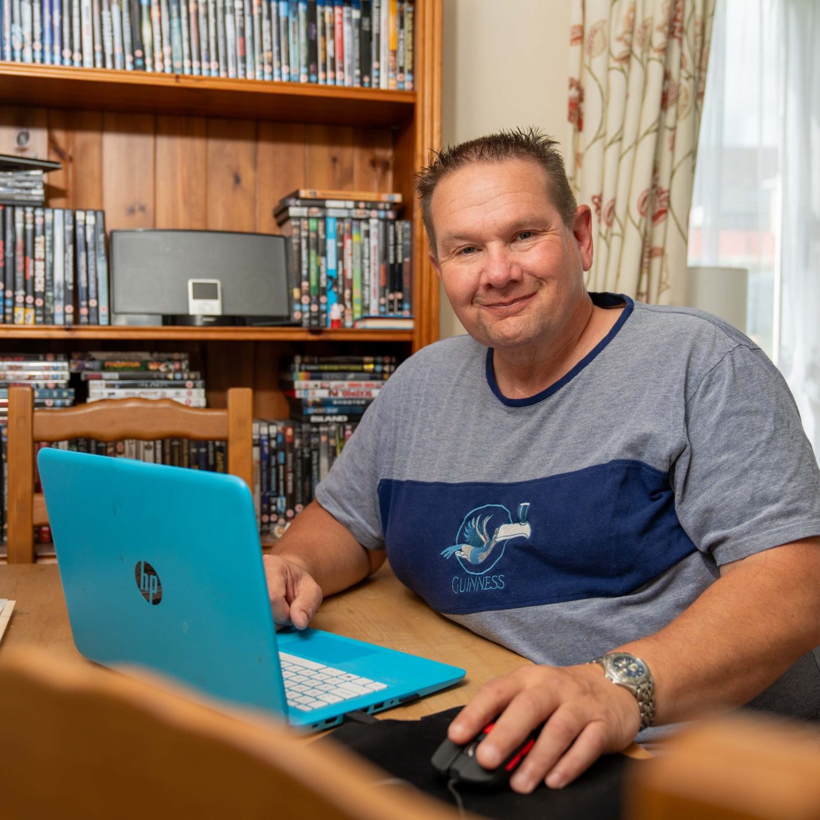 Trivallis Housing Landlord Wales A man seated at a wooden table with a blue laptop, smiling at the camera, with a bookshelf filled with DVDs in the background, inside his Trivallis housing in RCT.