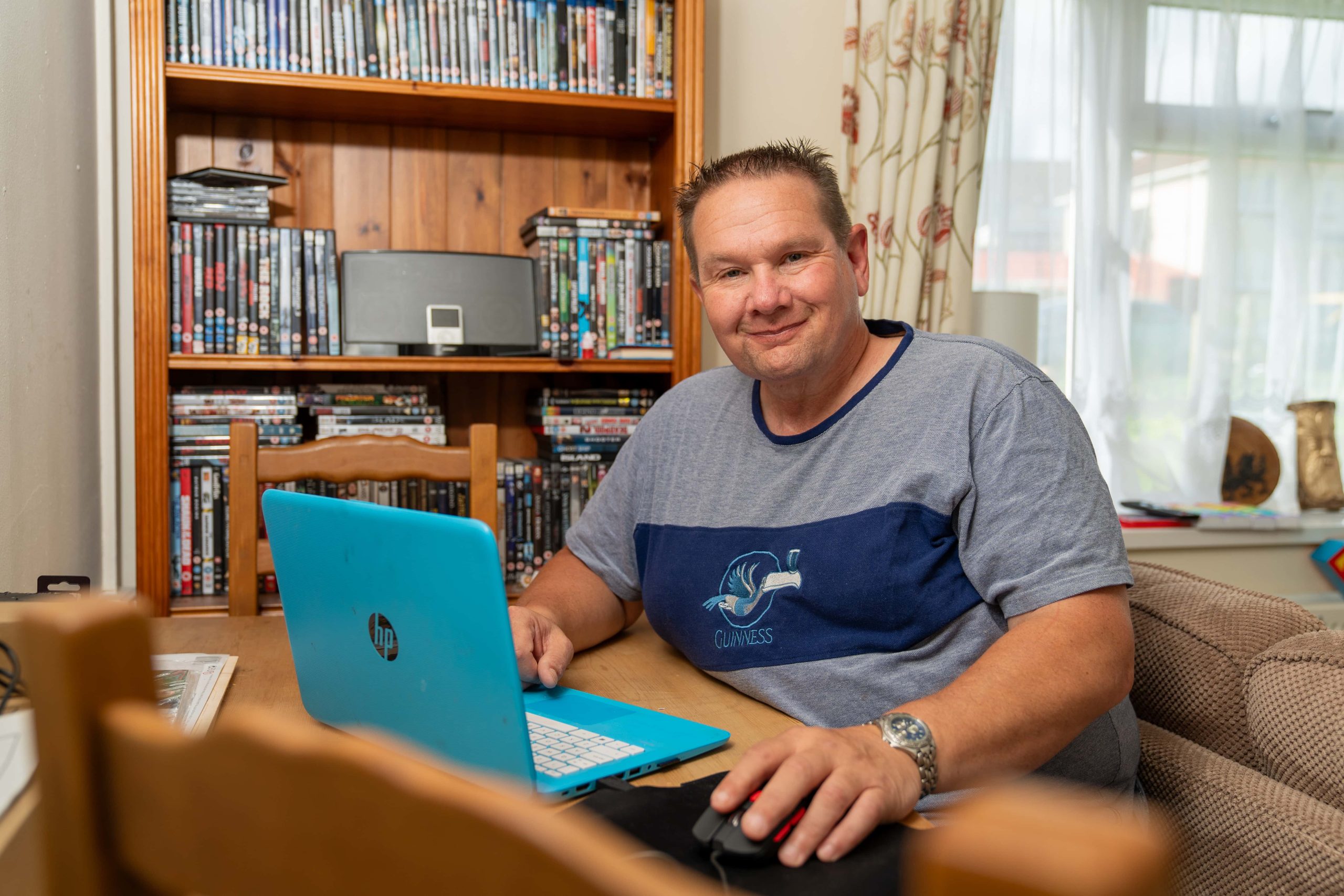 Trivallis Housing Landlord Wales A man seated at a wooden table with a blue laptop, smiling at the camera, with a bookshelf filled with DVDs in the background, inside his Trivallis housing in RCT.