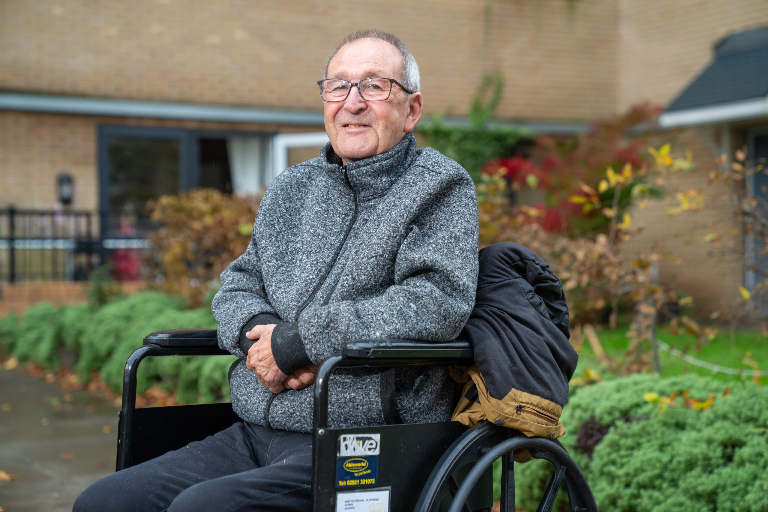 Trivallis Housing Landlord Wales An elderly man in a wheelchair is smiling while posing for the camera, with Trivallis housing and greenery in the background.
