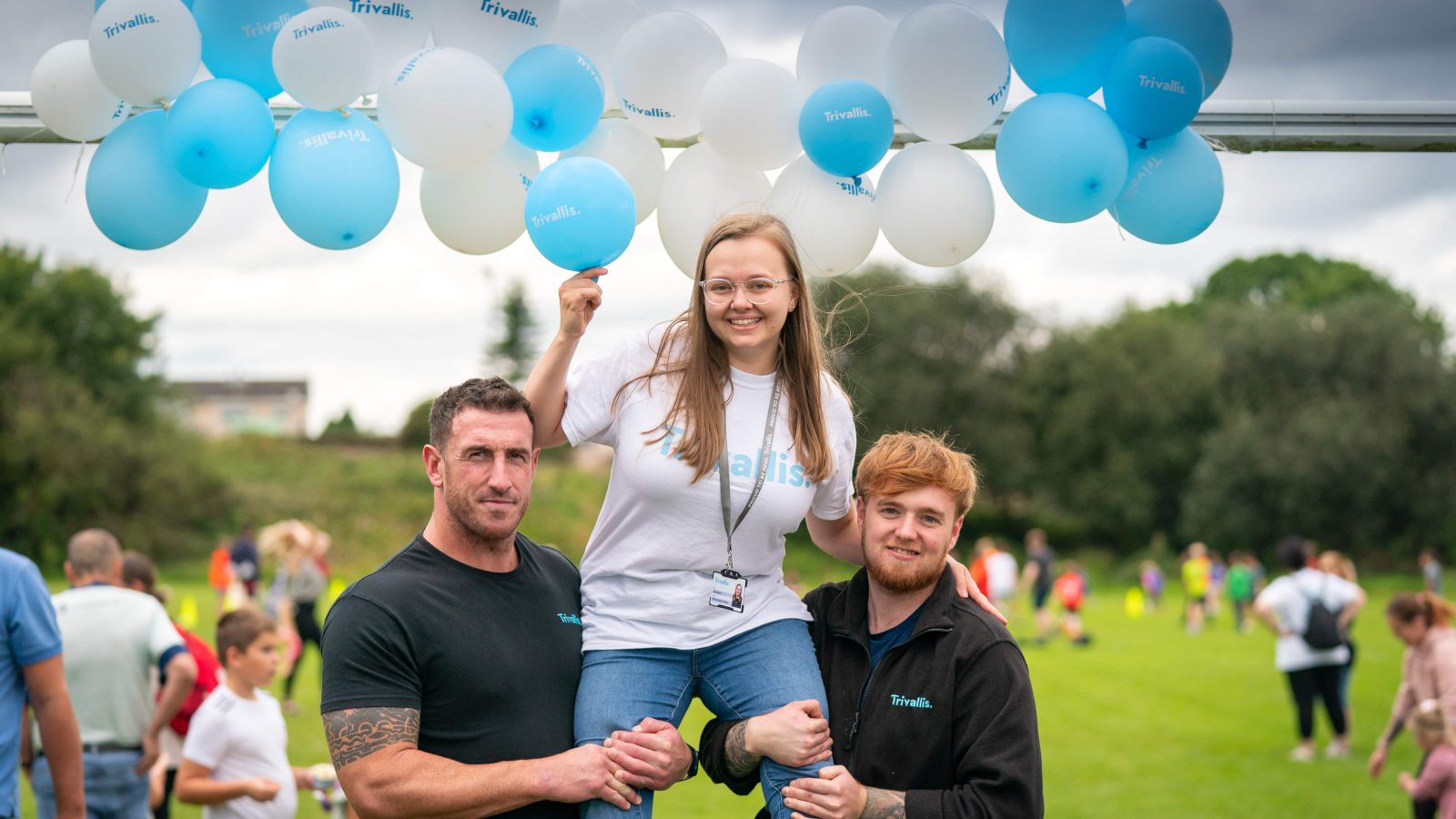 Trivallis Housing Landlord Wales Two men lifting a woman on their shoulders at an outdoor RCT housing event with blue and white balloons in the background.