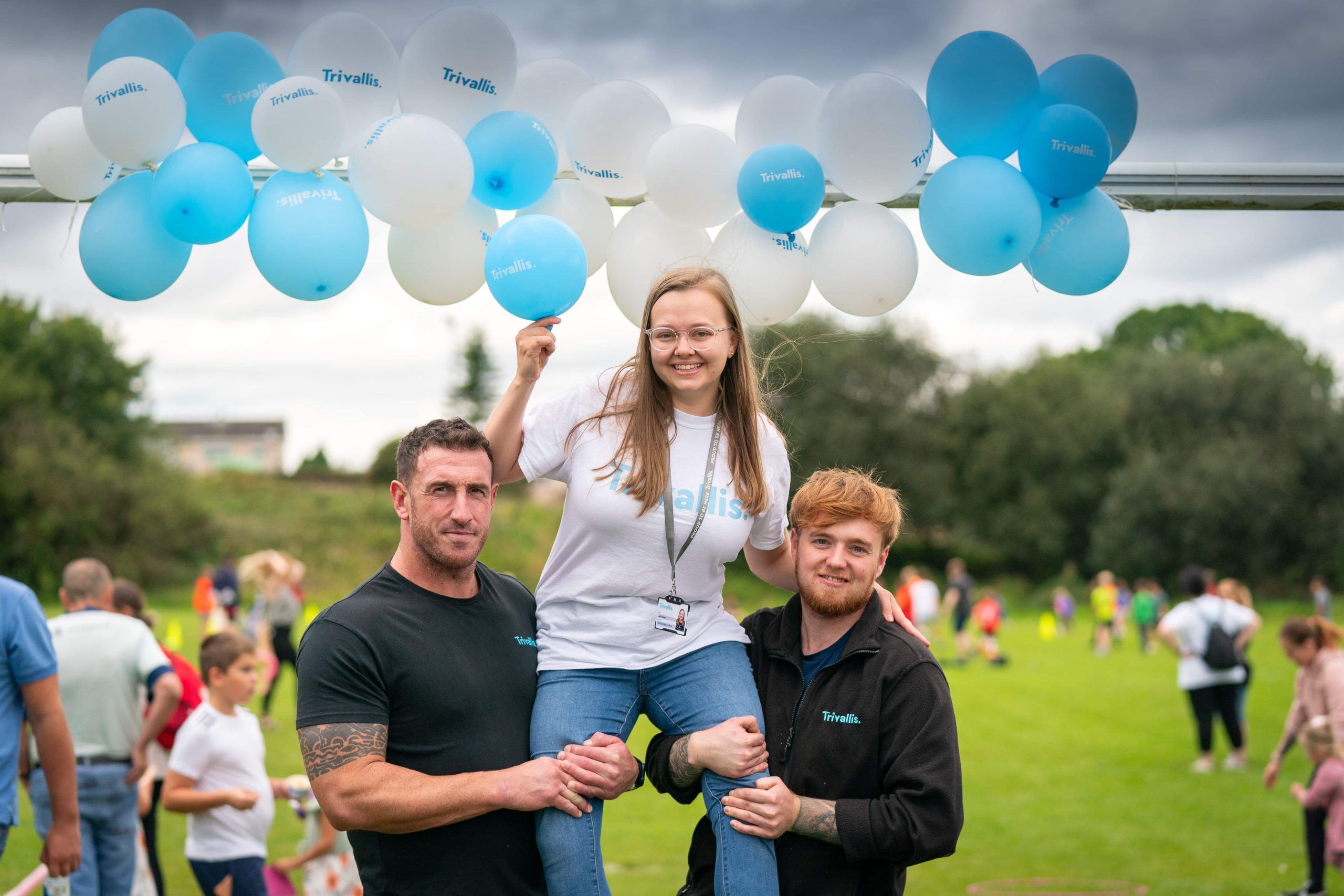 Trivallis Housing Landlord Wales Two men lifting a woman on their shoulders at an outdoor RCT housing event with blue and white balloons in the background.