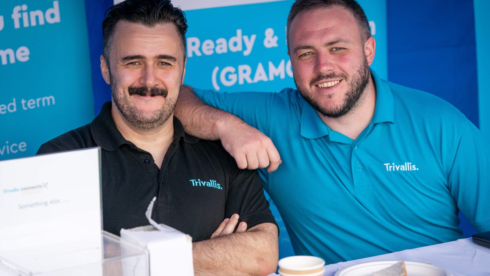 Trivallis Housing Landlord Wales Two men wearing Trivallis-branded polo shirts are smiling and posing at an outdoor housing event, with a promotional stand in the background and a table with paperwork and a coffee cup in the foreground.