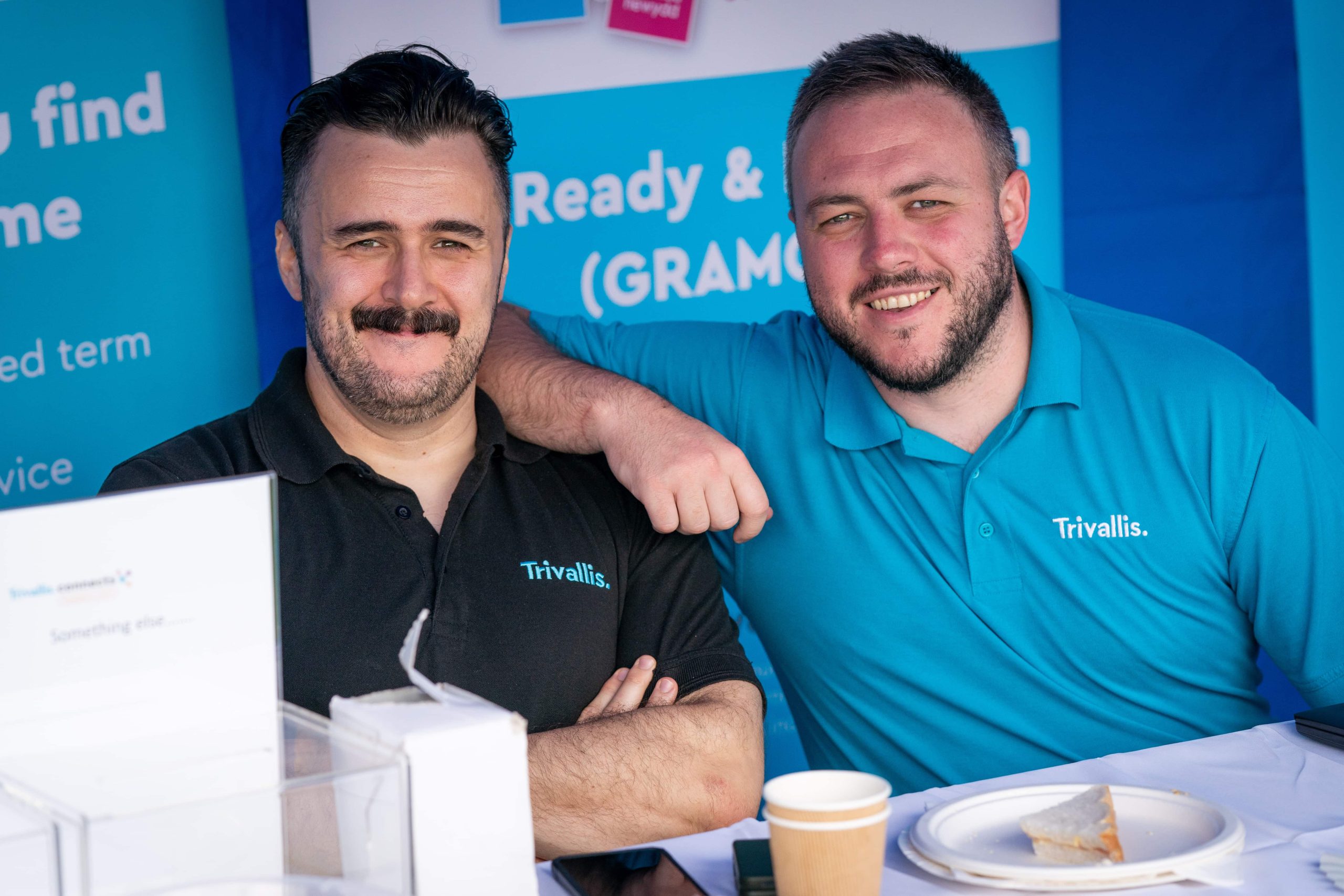 Trivallis Housing Landlord Wales Two men wearing Trivallis-branded polo shirts are smiling and posing at an outdoor housing event, with a promotional stand in the background and a table with paperwork and a coffee cup in the foreground.