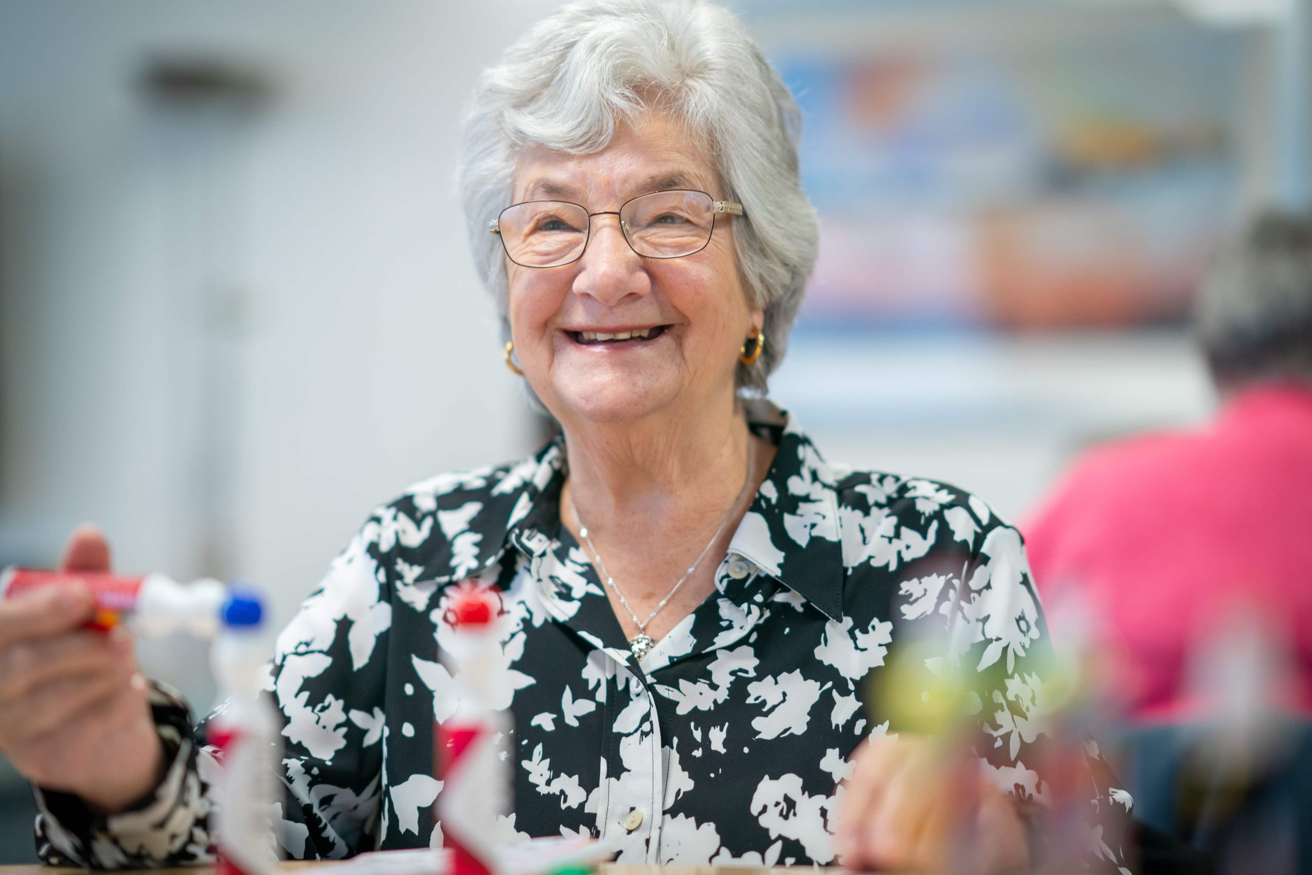 Trivallis Housing Landlord Wales An elderly woman with gray hair and glasses smiling while in a Trivallis housing room, with blurred figures in the background.