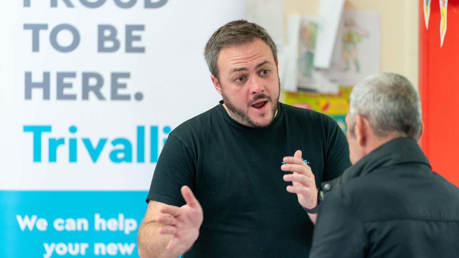 Trivallis Housing Landlord Wales Two men engaged in a conversation, with one man speaking and gesturing with his hands while the other listens, in front of a sign that says 