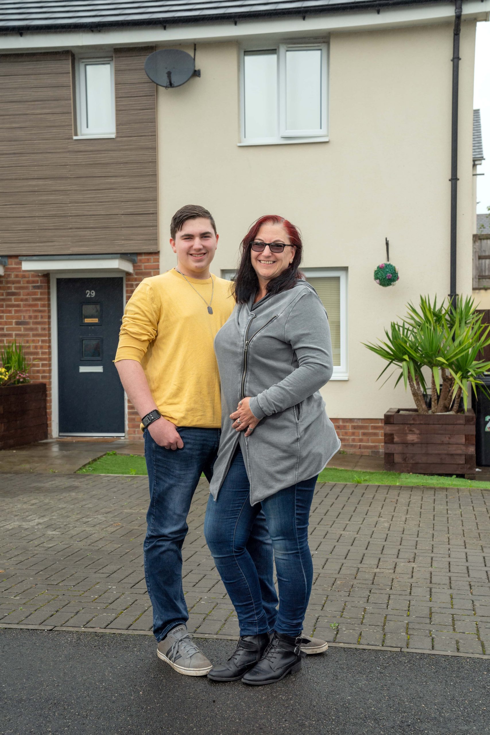 Trivallis Housing Landlord Wales A woman and a younger male companion, possibly her son, are standing together in front of a Trivallis housing property with the visible house number 29 on a cloudy day. Both are smiling and
