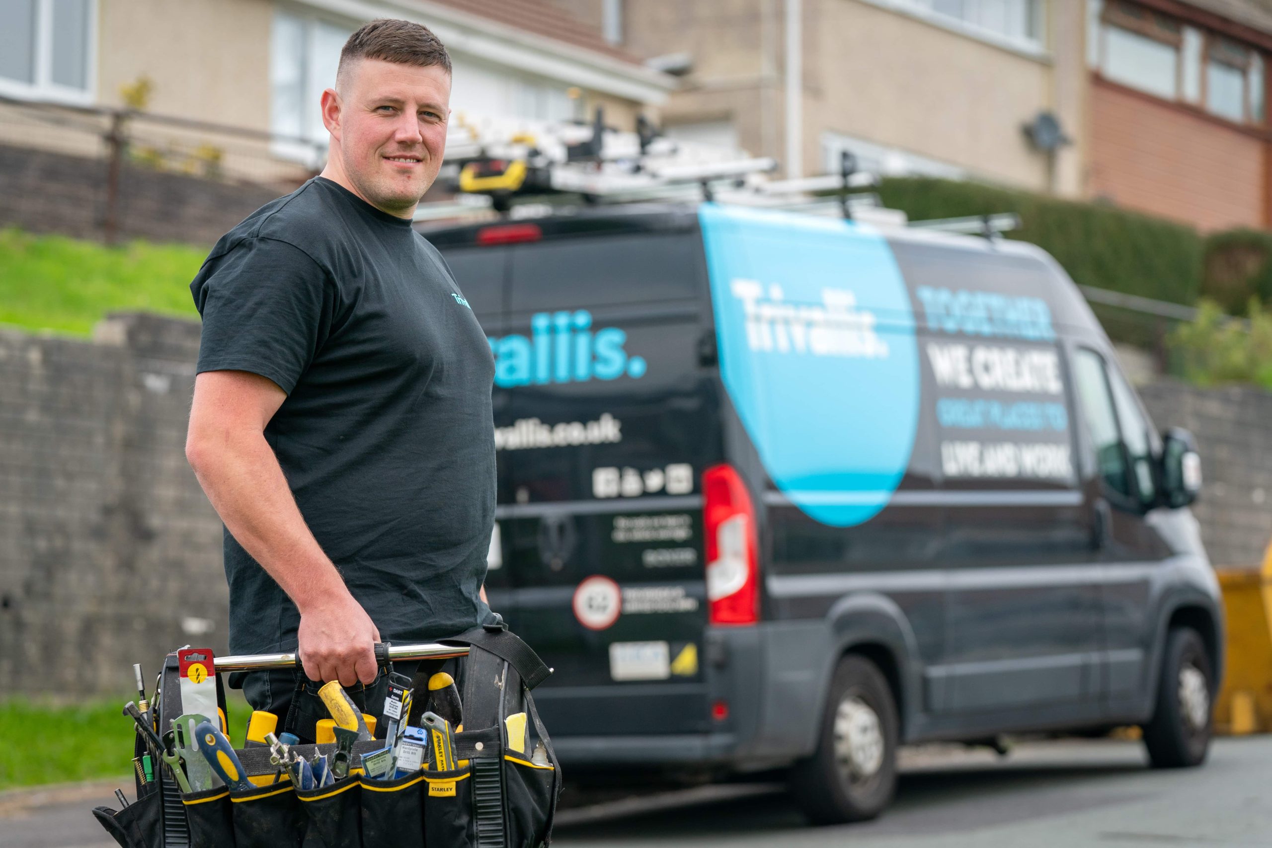 A man carrying a toolbox smiling in front of a service van with "trivallis" branding.
