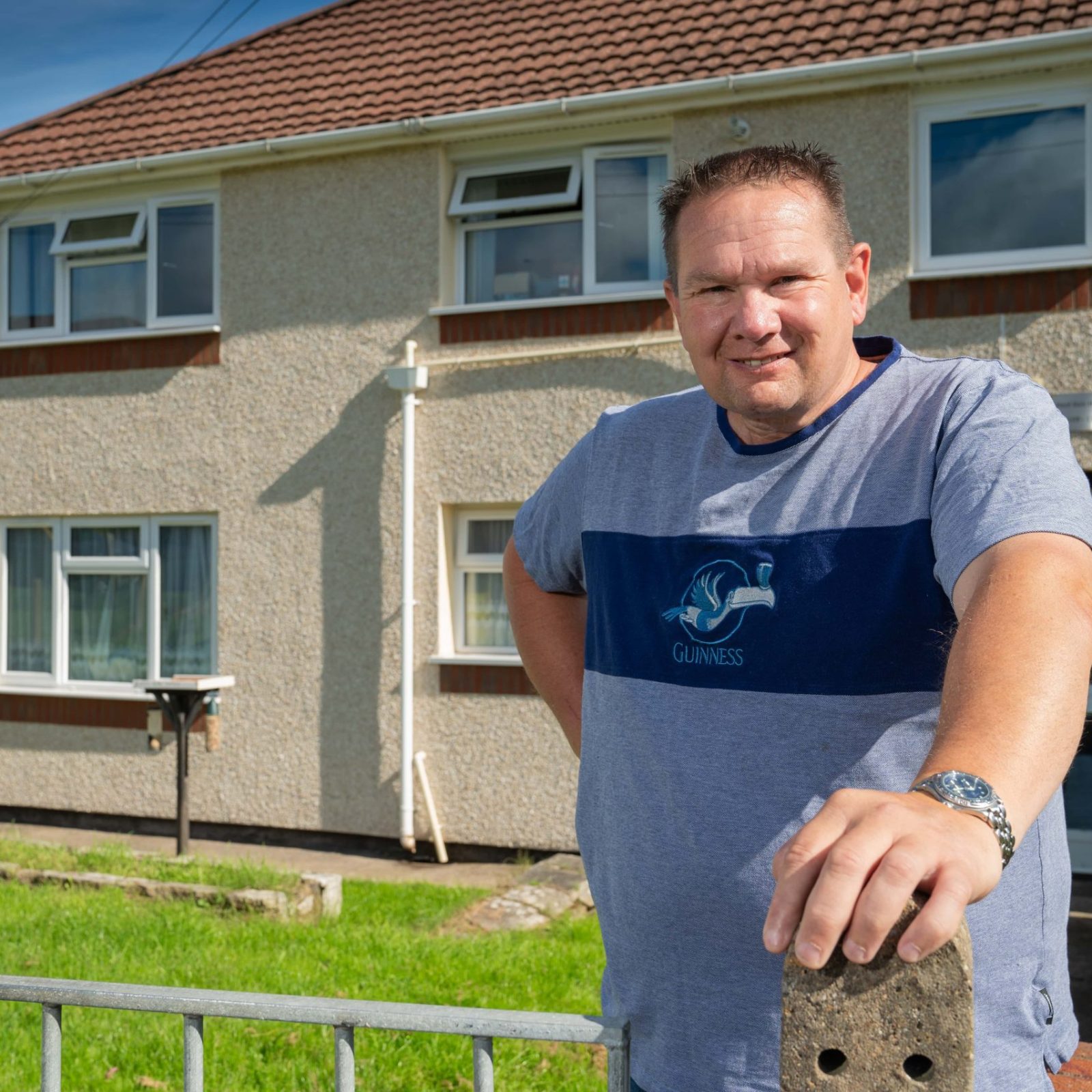 Trivallis Housing Landlord Wales A man stands in front of a two-story Trivallis house with a satellite dish on a sunny day, leaning on a metal fence with green grass in the background.