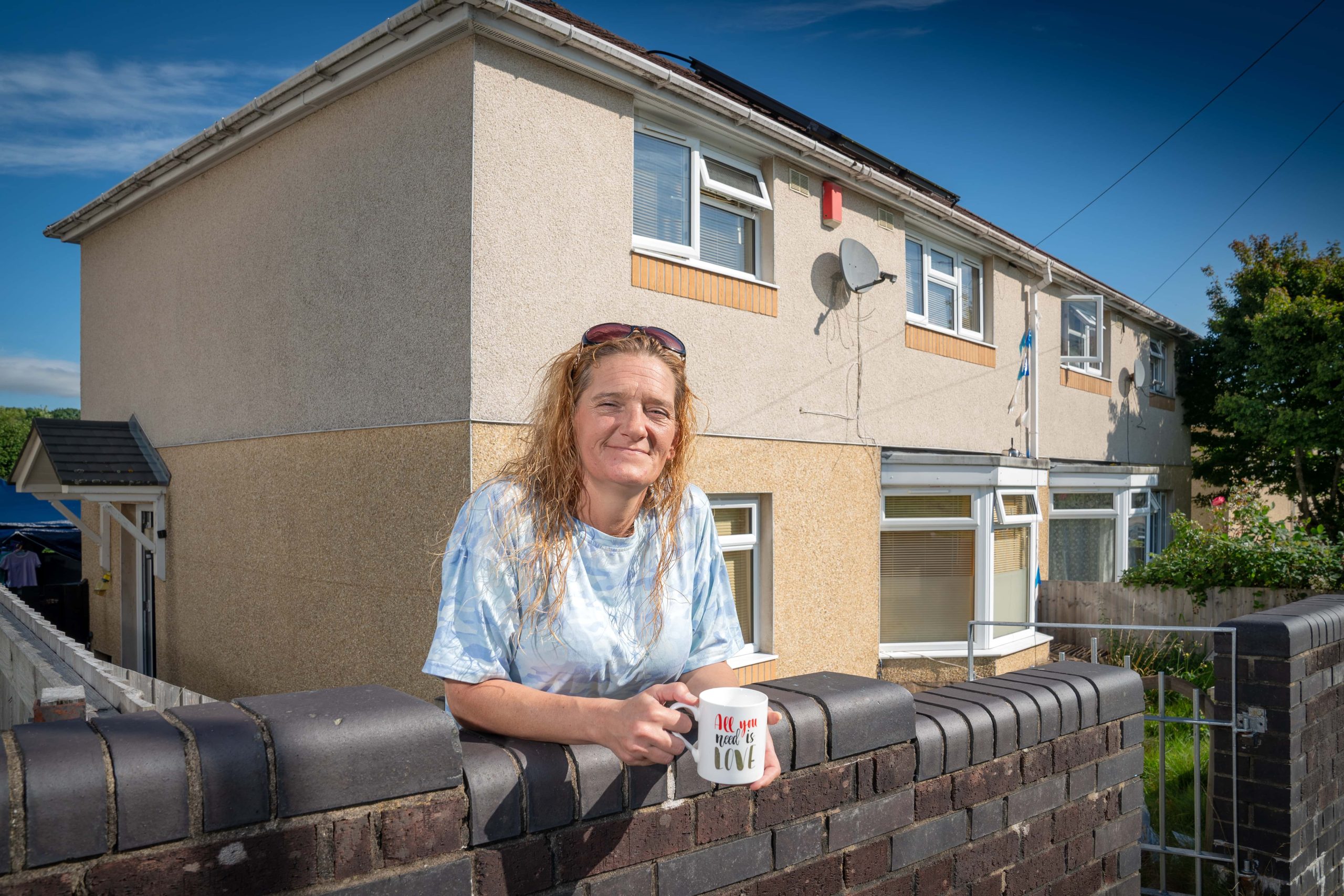 Trivallis Housing Landlord Wales A woman leaning on a brick fence in front of a Trivallis residential building, holding a mug and smiling at the camera.