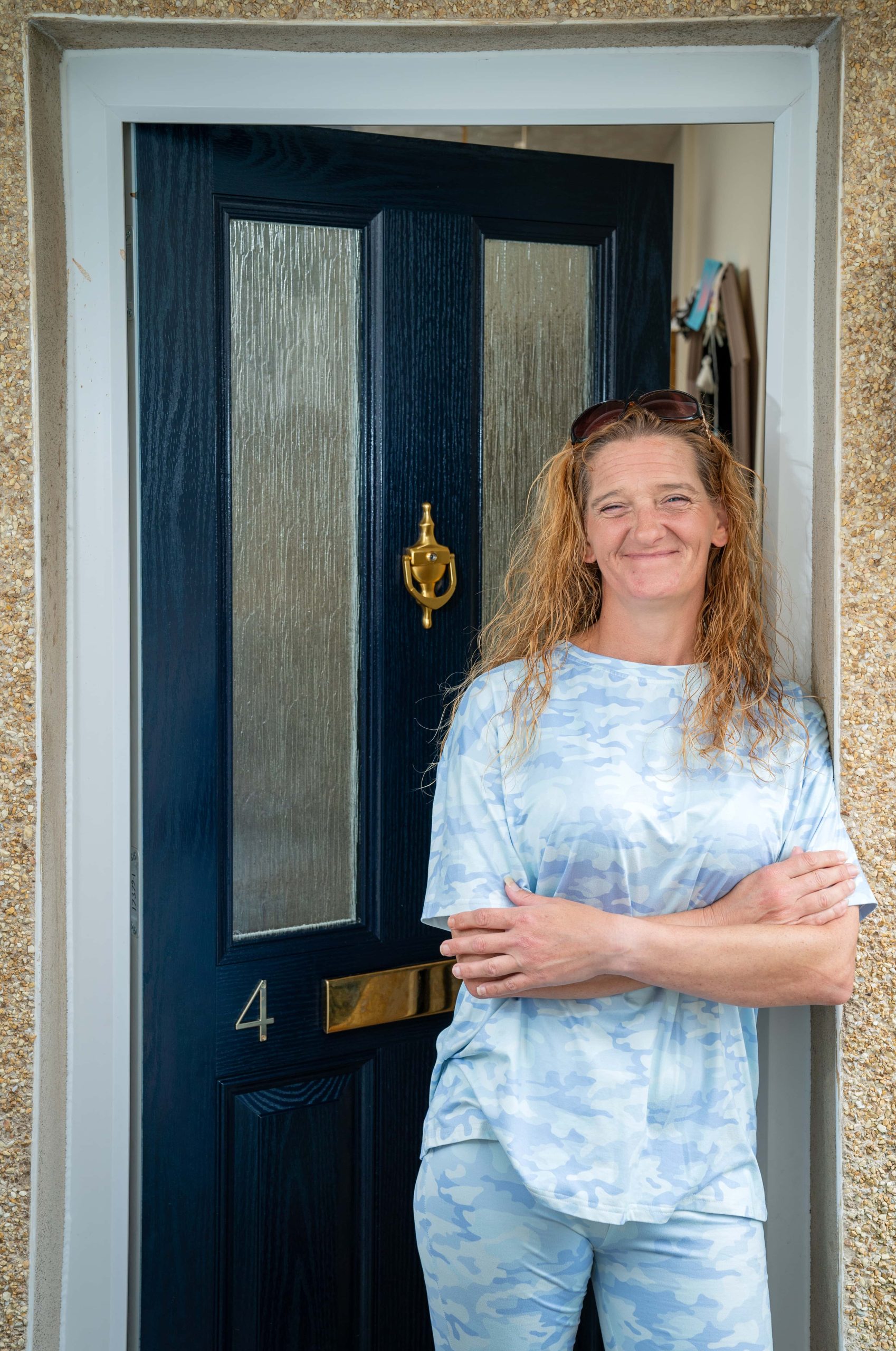 Trivallis Housing Landlord Wales A woman with long hair, wearing a blue and white patterned outfit, stands smiling with her arms crossed in front of a dark blue door marked with the number 4, emblematic of Triv