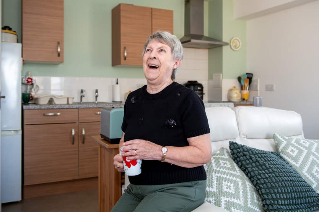 Trivallis Housing Landlord Wales An elderly woman is laughing while sitting on a couch in a well-lit, modern kitchen area in a Trivallis RCT housing unit, holding a red and white mug in her hand.