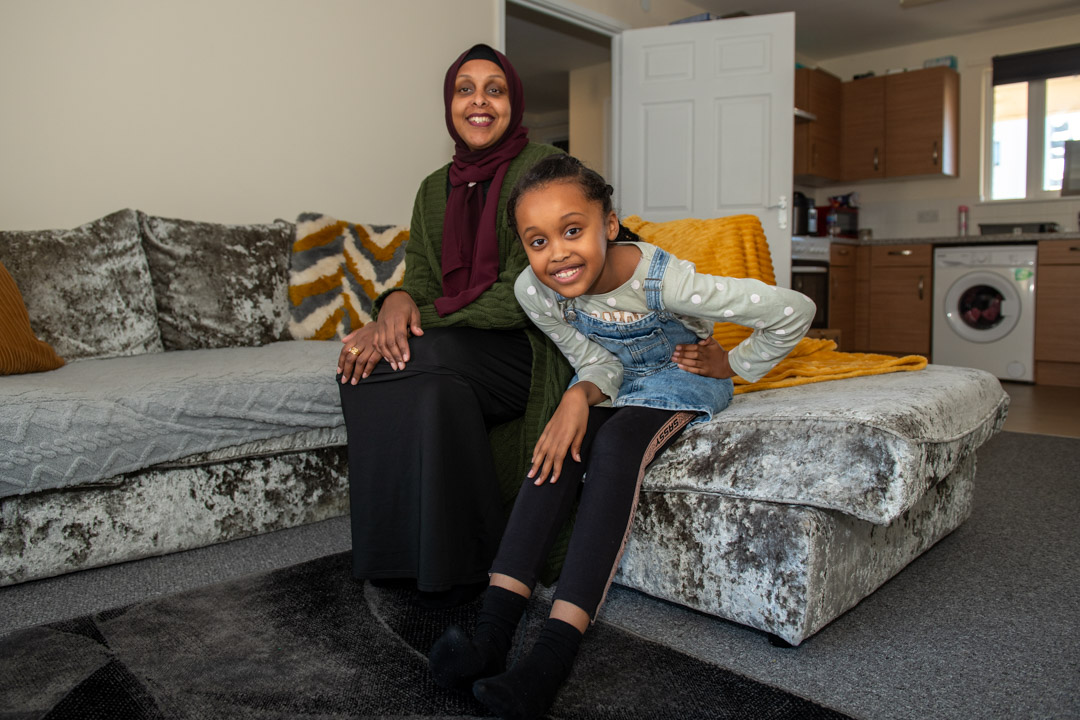 Trivallis Housing Landlord Wales A woman and a young girl are sitting in a Trivallis housing living room with a sofa and a kitchen area in the background. The woman is wearing a hijab and smiling at the camera,