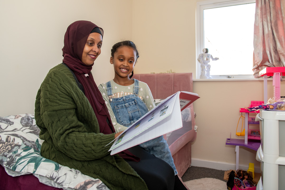 Trivallis Housing Landlord Wales A woman in a hijab and a young girl sitting on a sofa reading a book together in a well-lit Trivallis housing room.