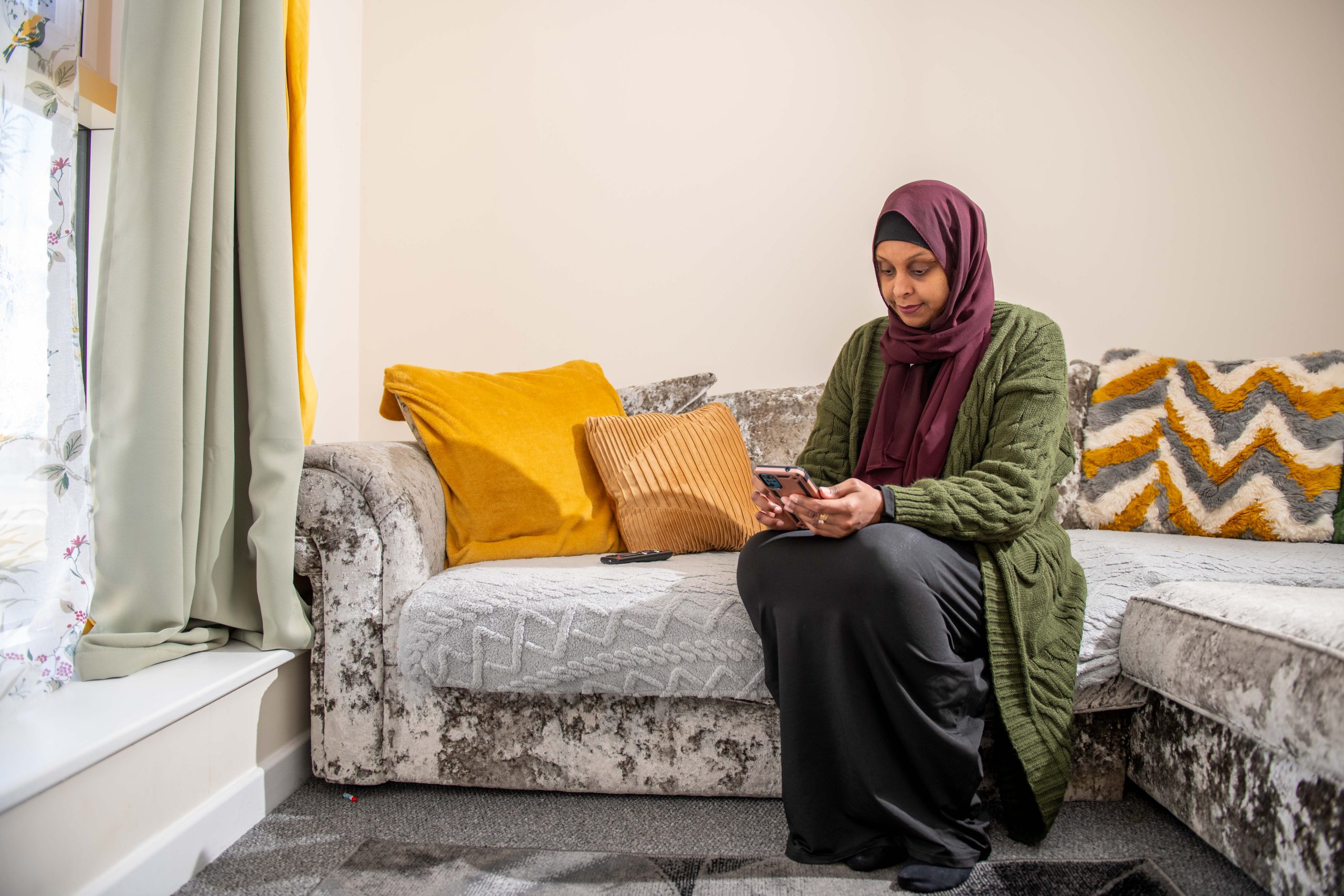 Trivallis Housing Landlord Wales A woman wearing a hijab sitting on a sofa and looking at her smartphone.