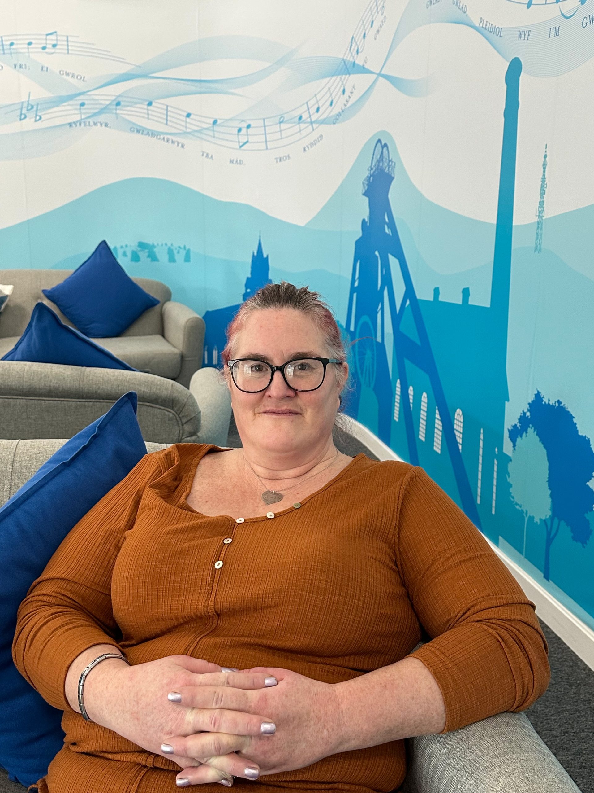 Trivallis Housing Landlord Wales A woman with glasses and a rust-colored top sitting comfortably on a grey couch with blue cushions, against a playful blue mural featuring musical notes and iconic structures.