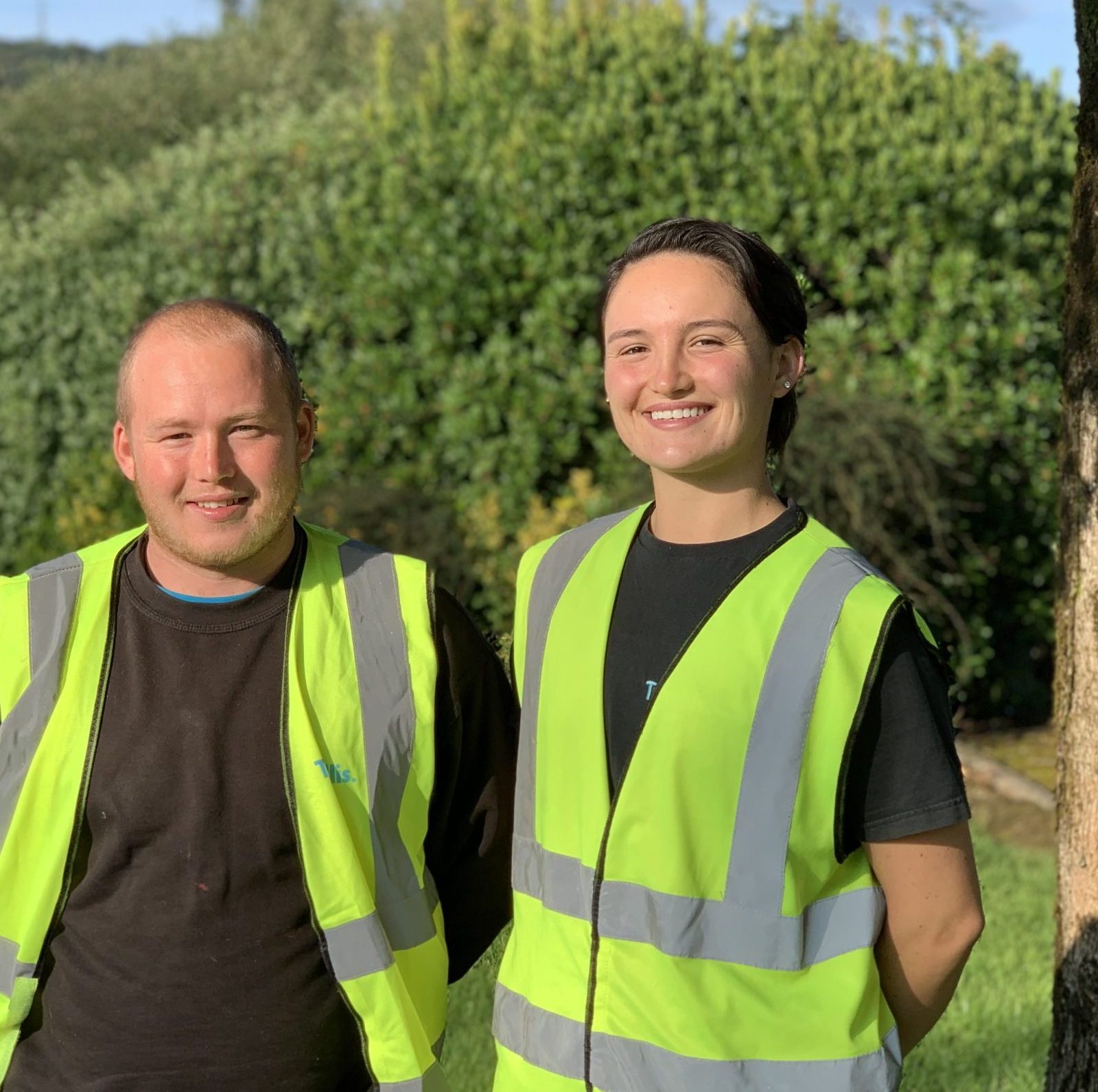 Trivallis Housing Landlord Wales Two workers doing apprenticeships, wearing high-visibility vests stand outdoors, smiling. the man is on the left and the woman is on the right, with a sunny green landscape and a tree in the background.