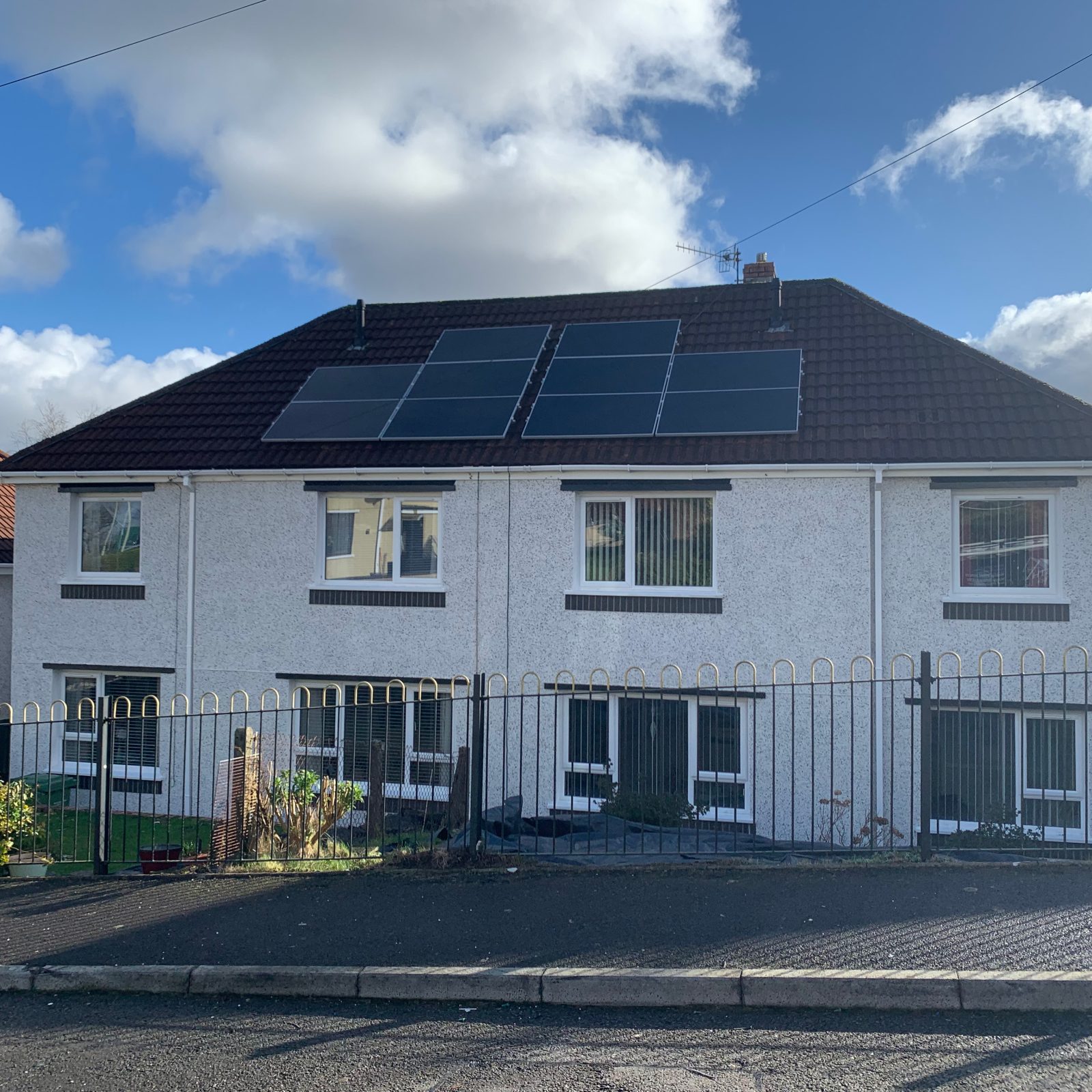 A two-story white pebbledash house with solar panels on the roof, black gates at the front, and a blue sky with scattered clouds above.