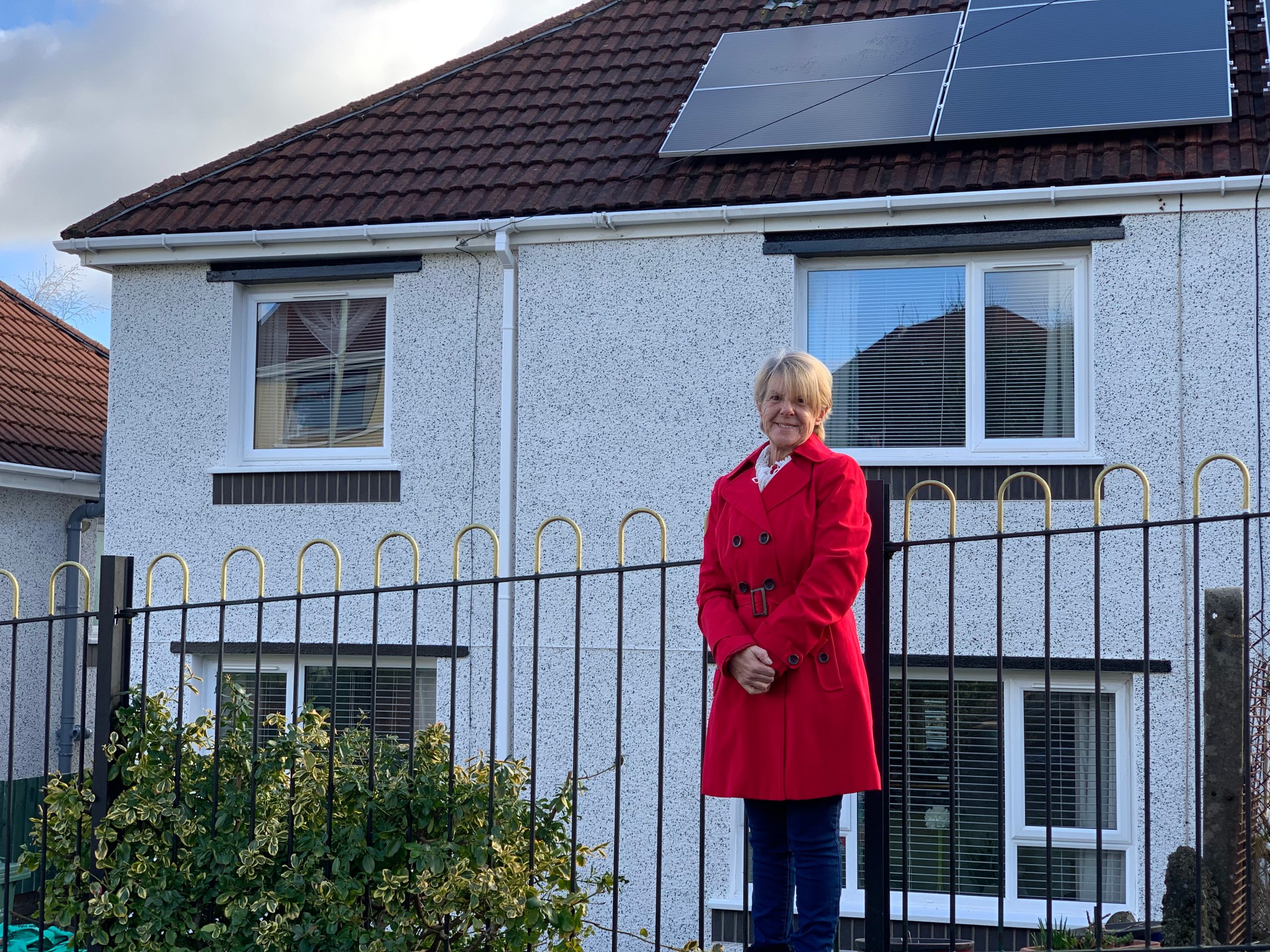 Trivallis Housing Landlord Wales A woman in a red coat standing in front of a white two-story house with solar panels on the roof.