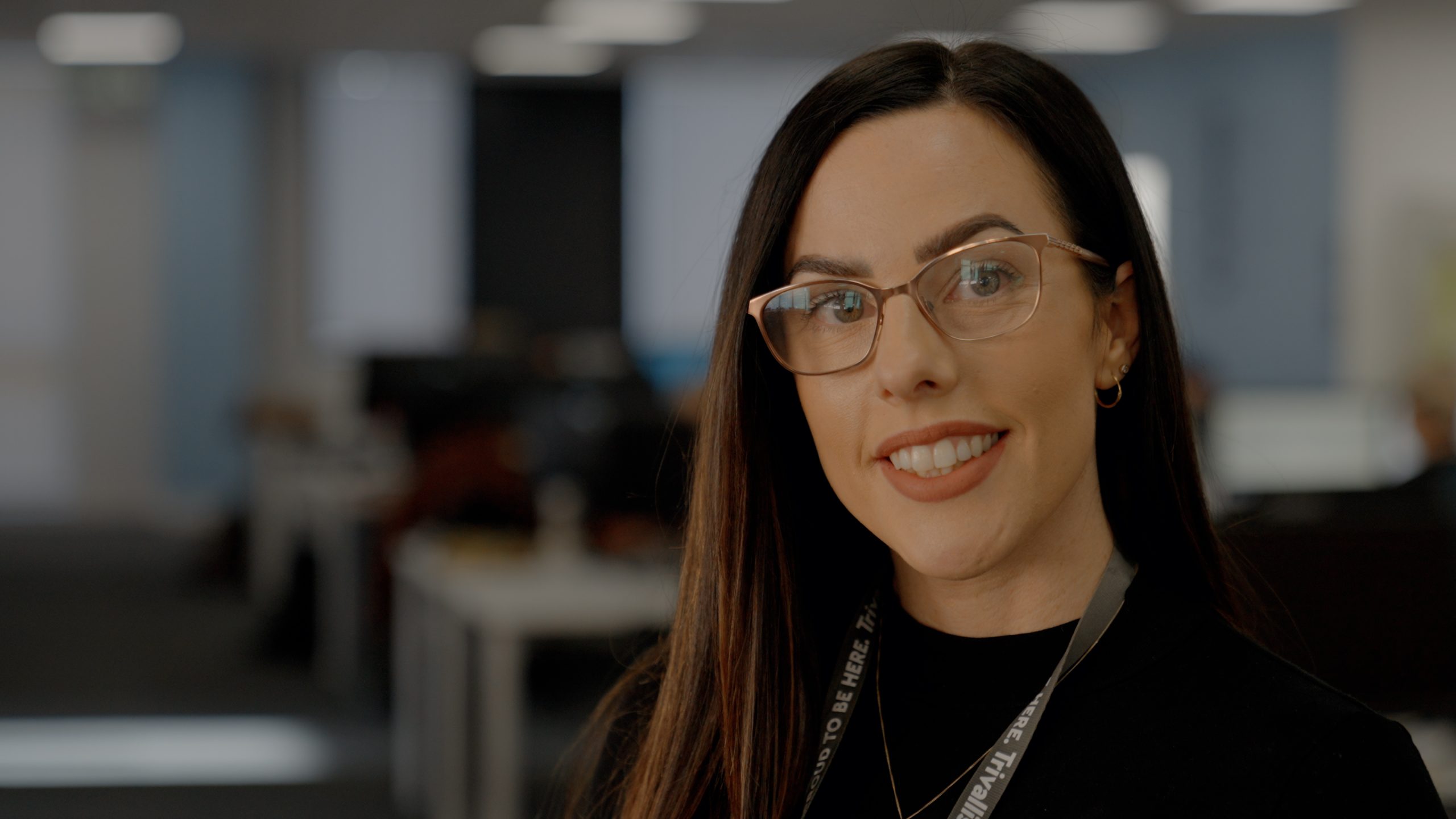 A professional woman with long dark hair and glasses is smiling at the camera. she is wearing a black turtleneck and a lanyard. the background shows a blurred office space with desks and computers.