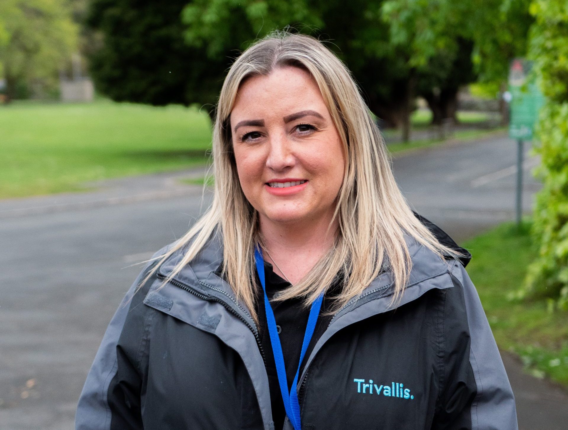 Trivallis Housing Landlord Wales A woman with long blonde hair stands outdoors, wearing a black jacket labeled "trivallis" and an id badge. a grassy area and trees are in the background.