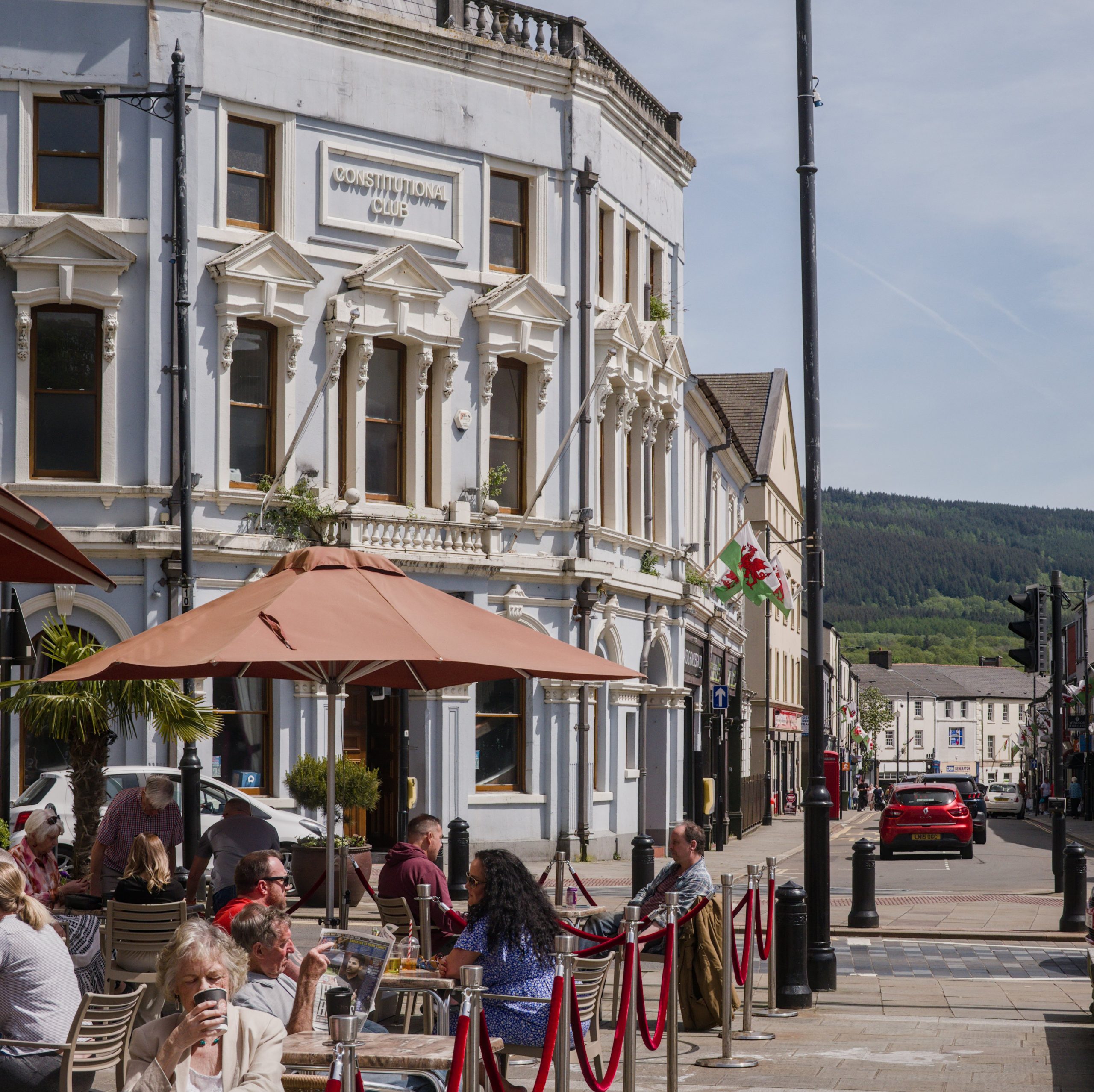 Trivallis Housing Landlord Wales Aberdare street scene featuring people dining outdoors at a cafe. The establishment is set in a light-coloured, ornate building labeled "Constitution Club." The street is lined with various shops, a few cars are parked, and hills are visible in the background.