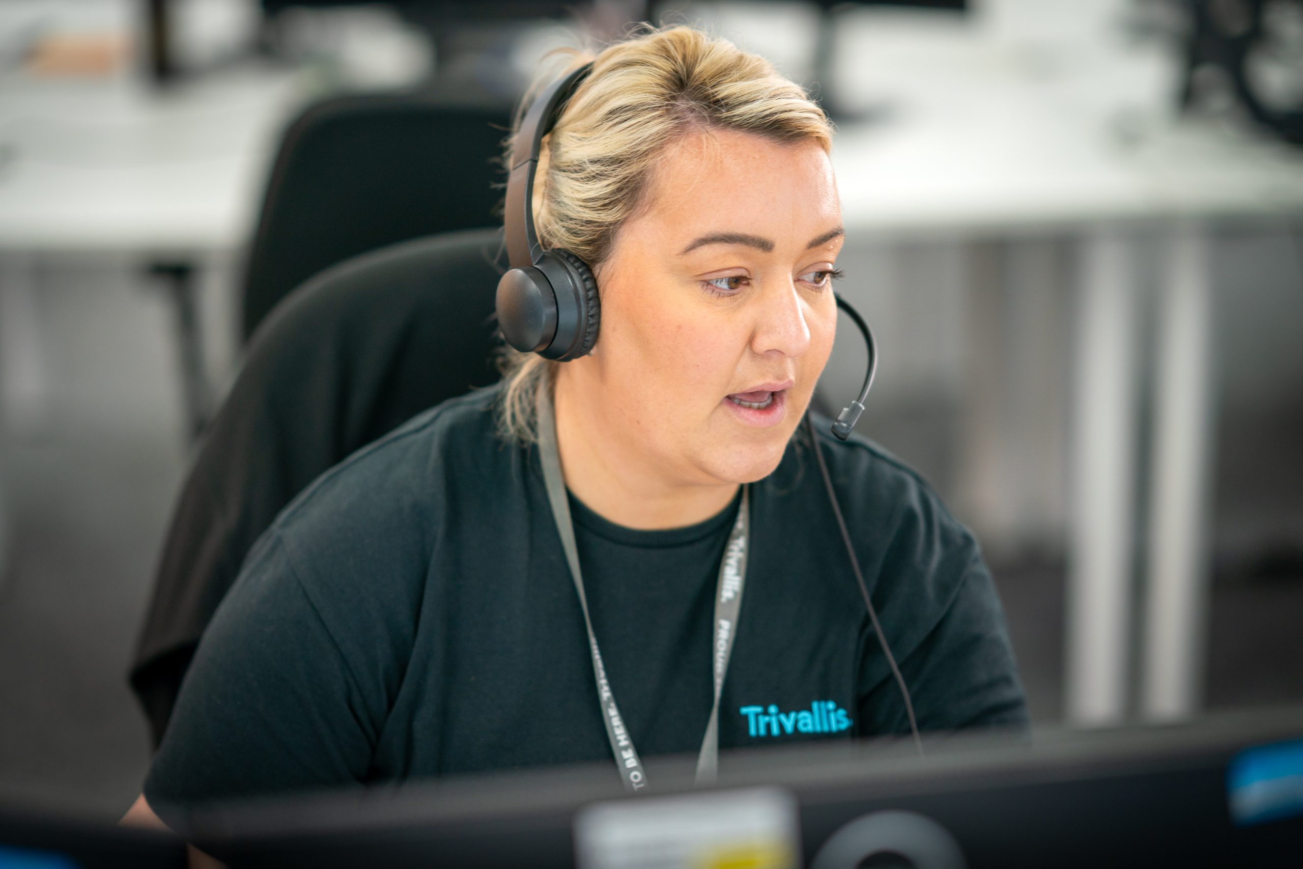 Trivallis Housing Landlord Wales A woman with short blonde hair, wearing a black t-shirt and headset, working at a computer in an office setting. she appears focused on her task. the name "trivallis" is visible on her shirt.