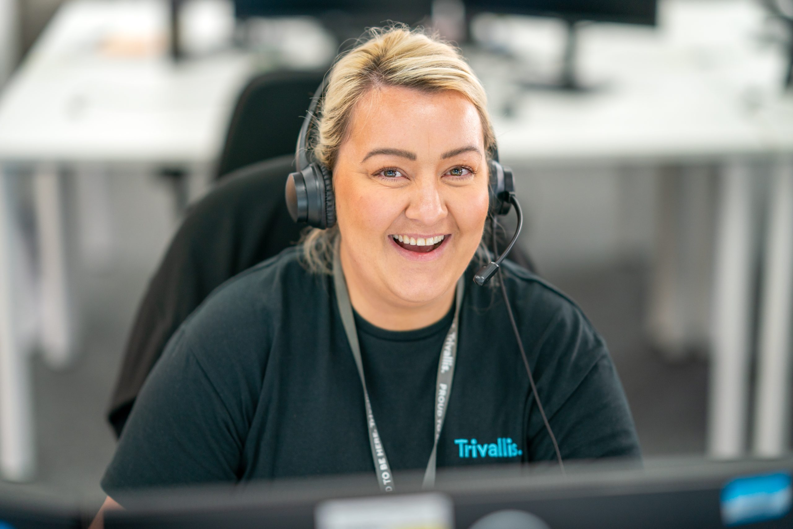 Trivallis Housing Landlord Wales A cheerful woman with blonde hair wearing a headset and a black shirt labelled "Trivallis" is sitting at her desk in a modern office, looking at a computer screen, apparently engaged in a customer service interaction.