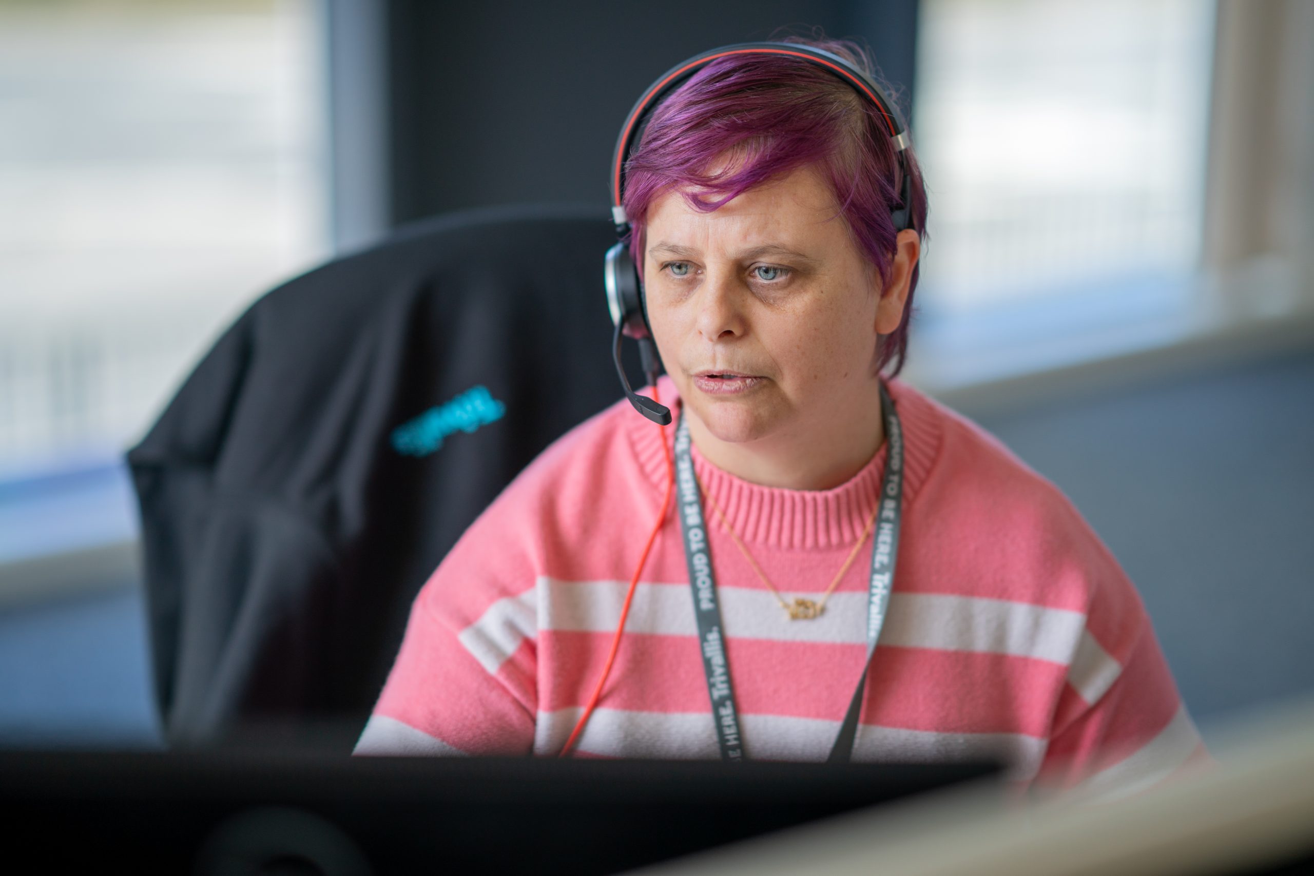 Trivallis Housing Landlord Wales A woman with purple hair wearing a headset sits in front of a computer monitor in an office setting. she is wearing a pink and white striped sweater and has a focused expression.