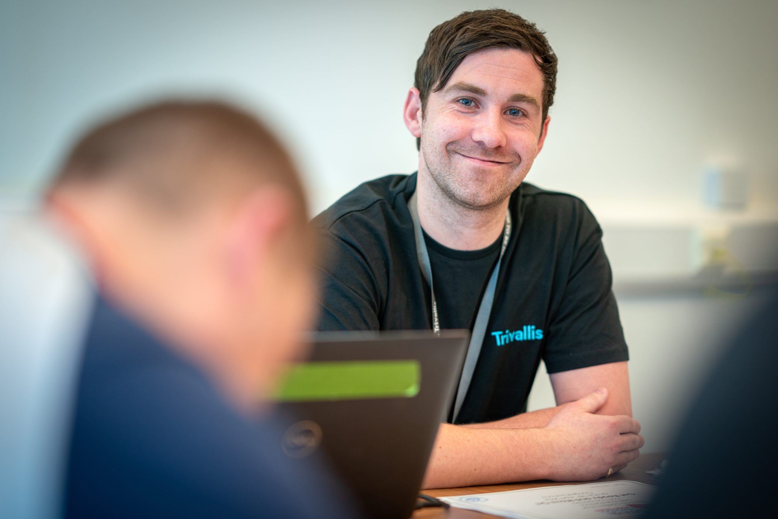 Trivallis Housing Landlord Wales A smiling man wearing a black t-shirt with the logo "trivallis" sits at a desk in an office, looking at the camera. a blurred colleague is visible in the foreground, facing a computer screen.