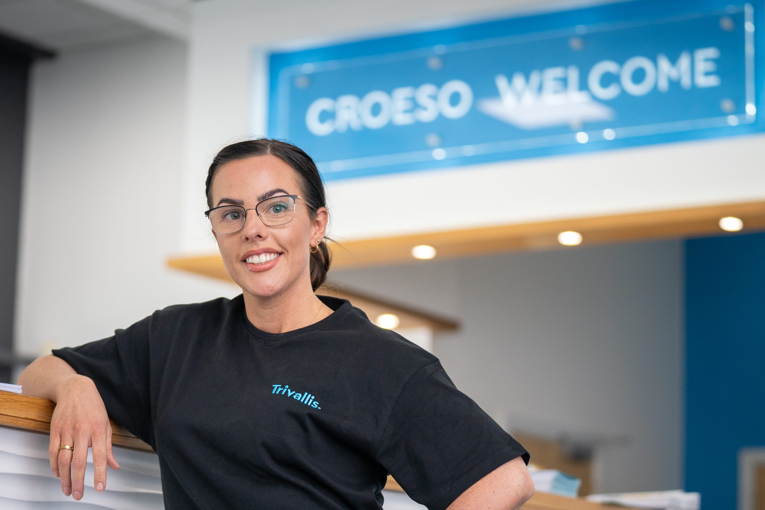 Trivallis Housing Landlord Wales A woman with glasses and a black shirt marked "trivallis" stands smiling at a reception desk. Above her, a sign reads "croeso welcome" in blue letters.