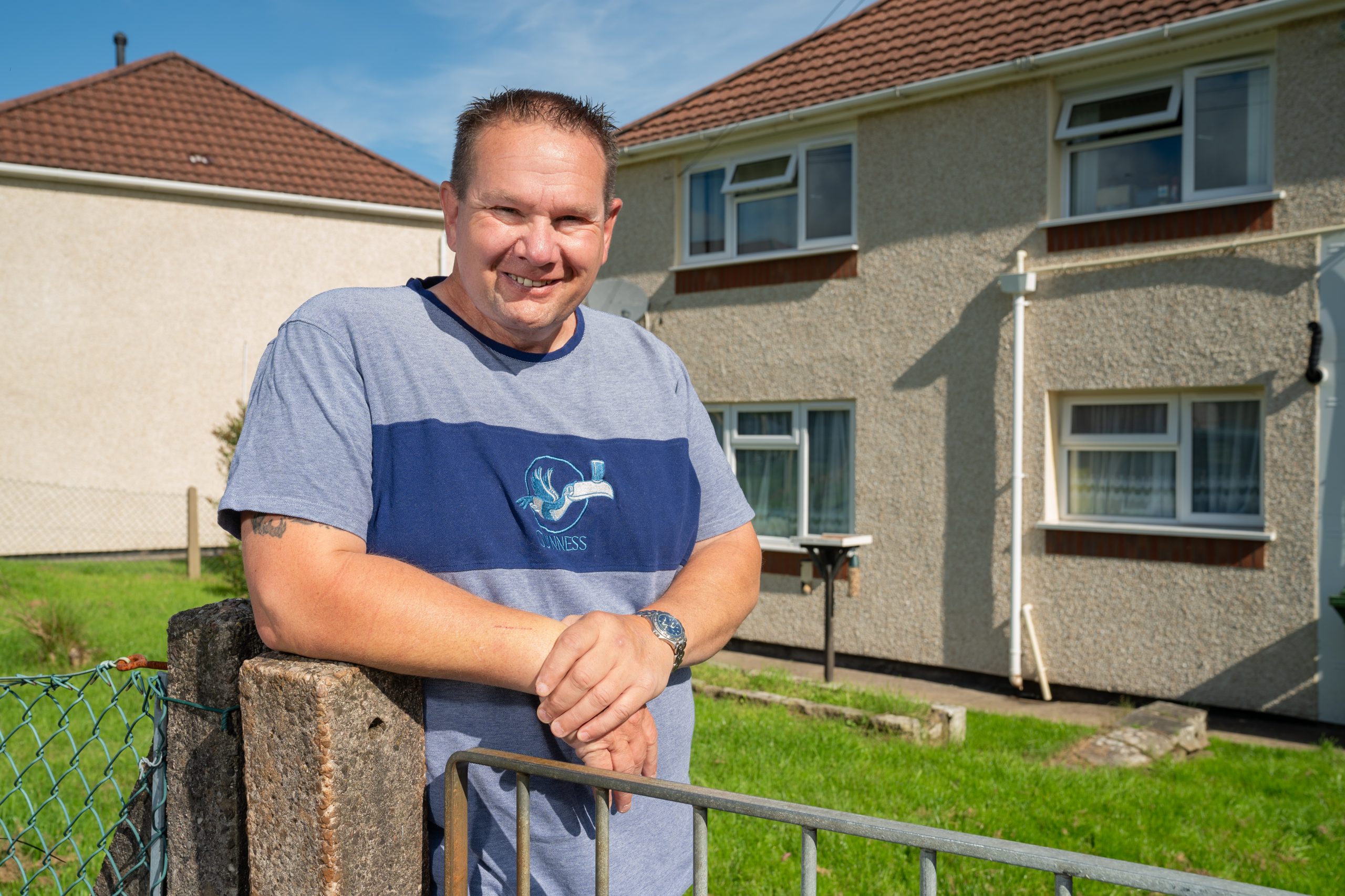 A smiling man leans on a metal gate in front of a suburban house with a well-kept lawn on a sunny day. he is wearing a blue t-shirt with a logo and casual shorts.